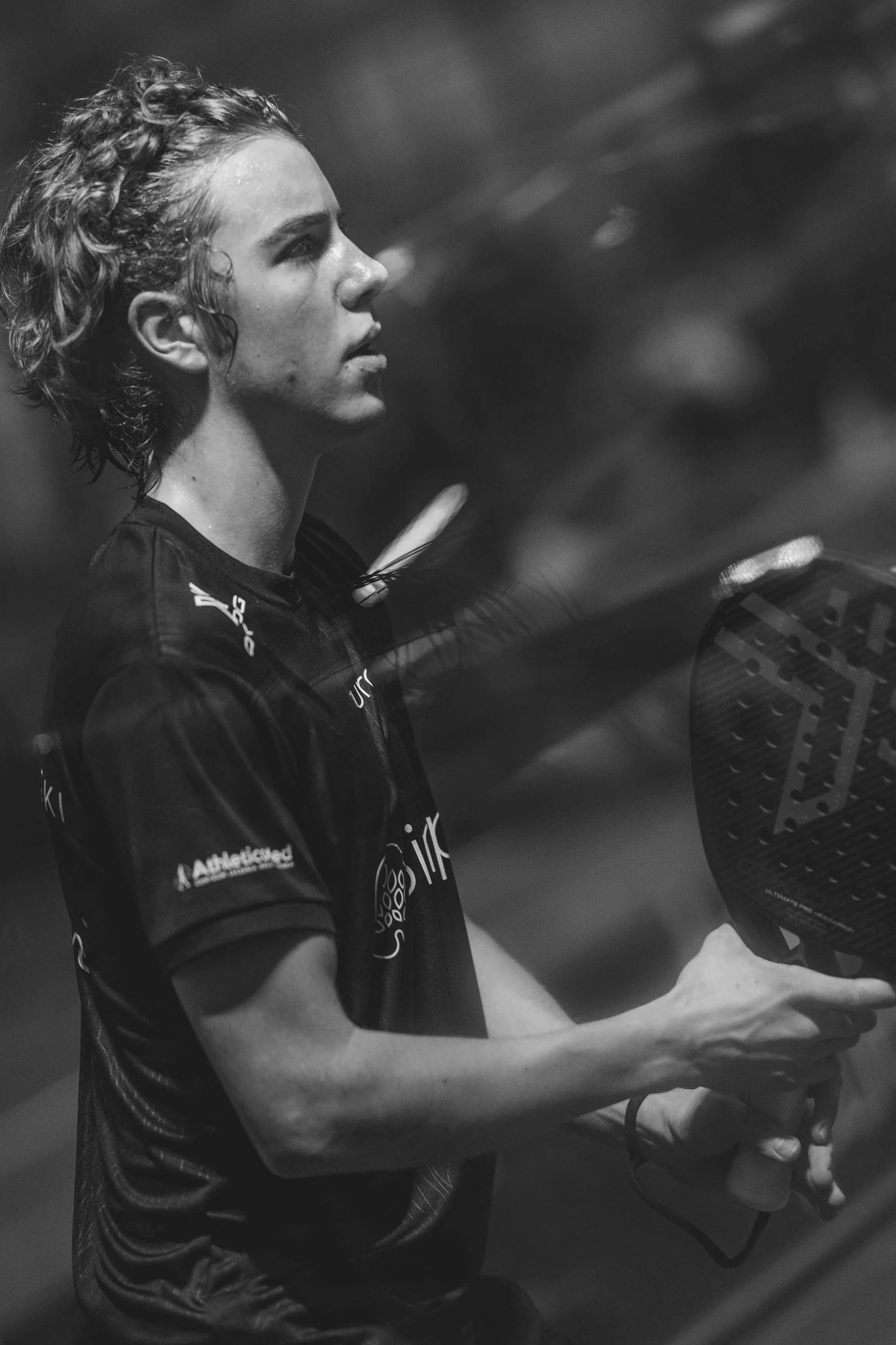 A woman with curly hair, wearing a sports shirt with logos, is playing pickleball with a paddle.