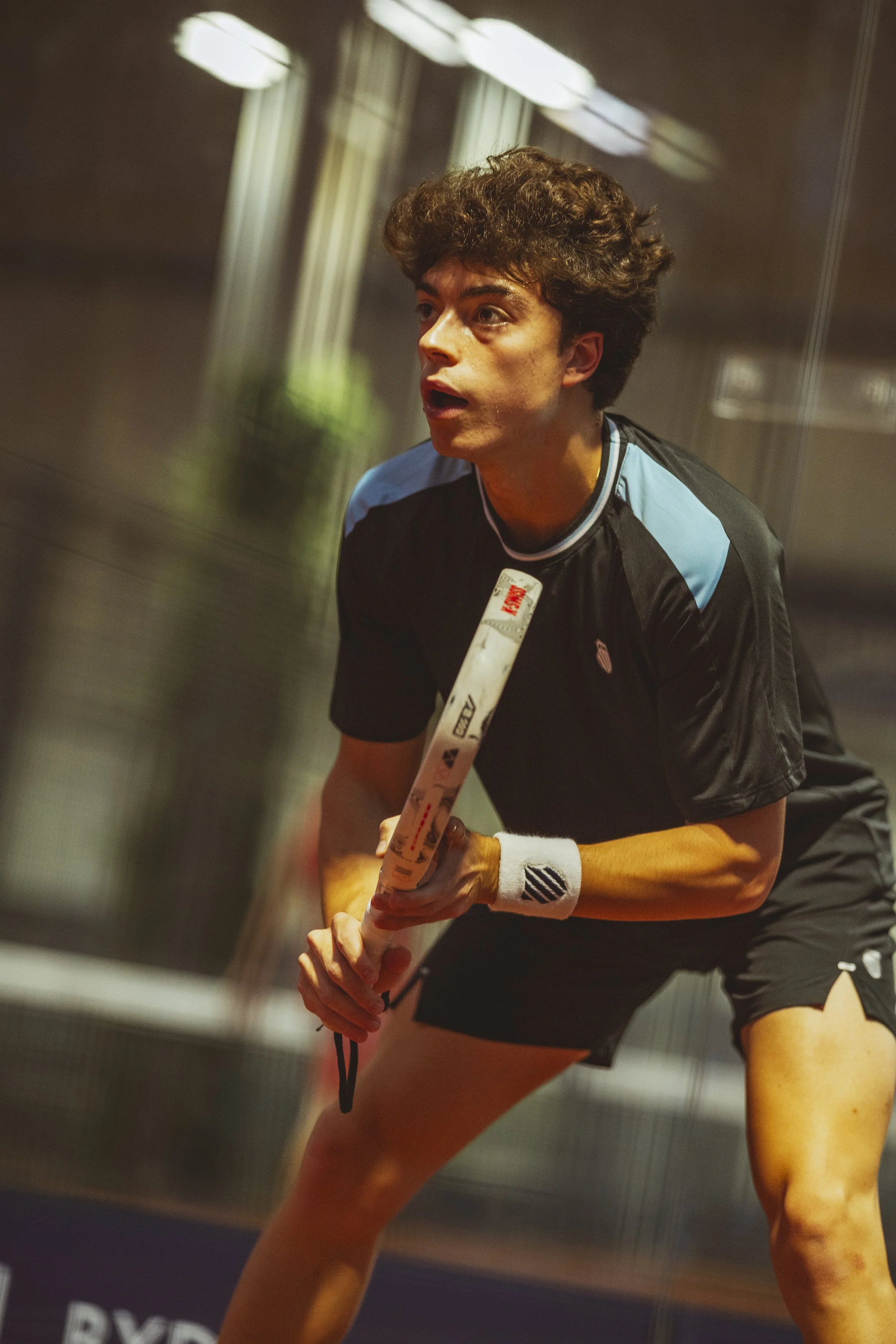 A young male athlete with curly brown hair wearing a black and light blue sports jersey, white wristband, and holding a hockey stick, appears focused and ready to play in an indoor sports facility.