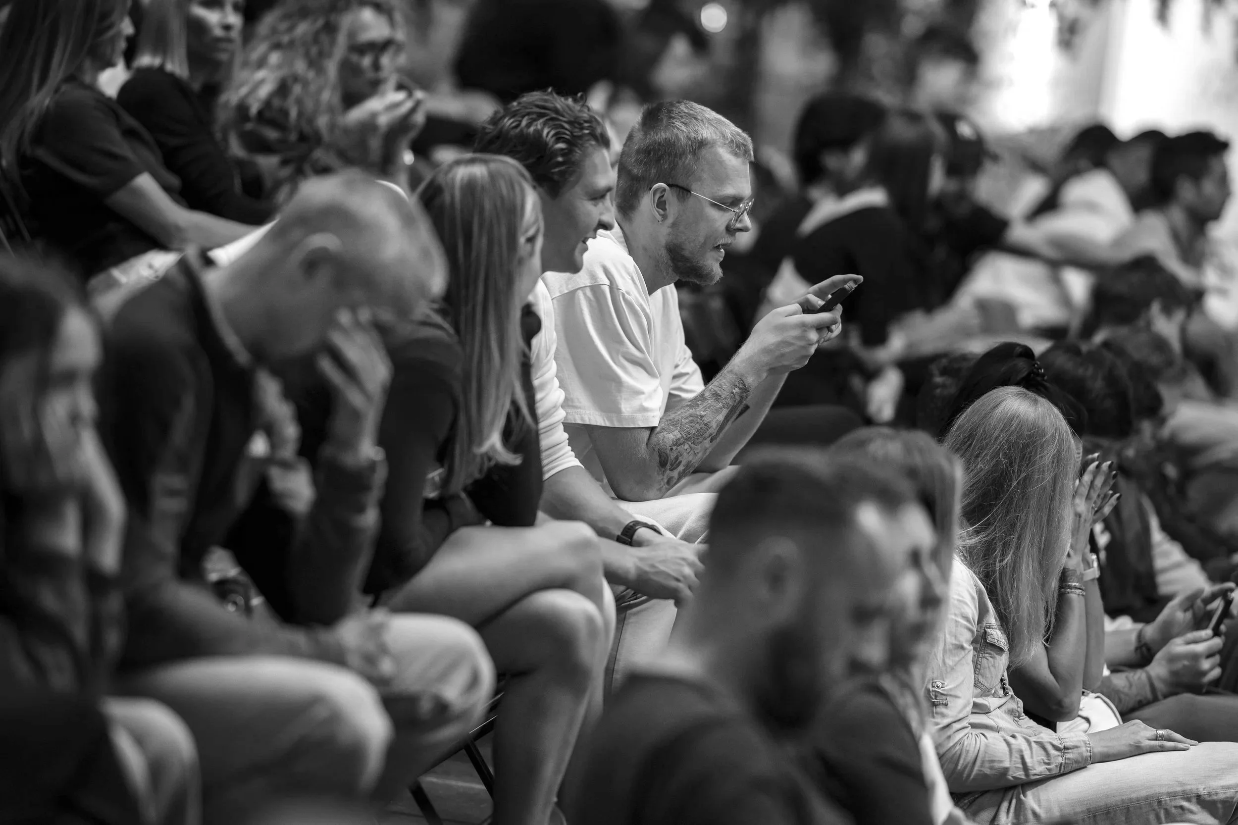 Black and white photo of a diverse group of people sitting in an audience, with some looking at their phones, watching or listening attentively.