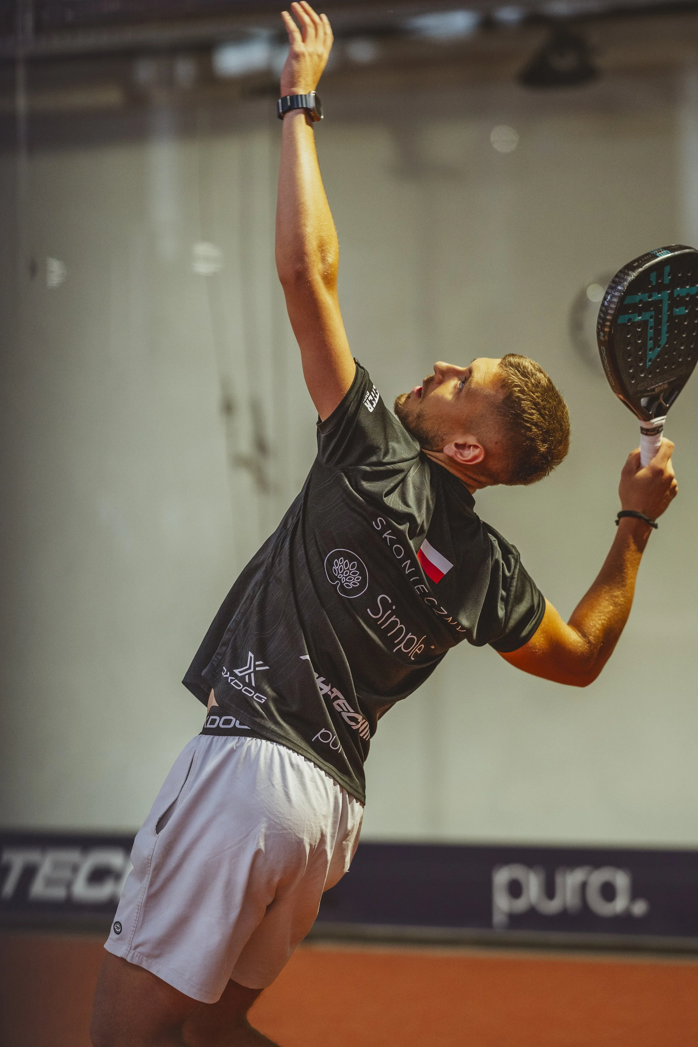 A male tennis player is serving on an indoor tennis court, wearing a black sports shirt with sponsor logos and white shorts, holding a tennis racket with a black handle and colorful strings, with his right arm extended upward.