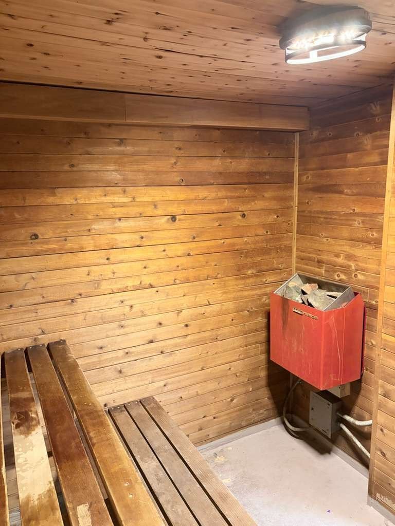 Sauna room with wood paneling on the walls and ceiling, a wooden bench on the left, and an electric heater with rocks in a red box on the right.