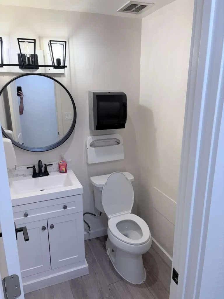 A small bathroom with a white vanity, round mirror, black faucet, hand soap dispenser, and a white toilet. There is a black paper towel dispenser above the toilet and a wall vent in the ceiling.