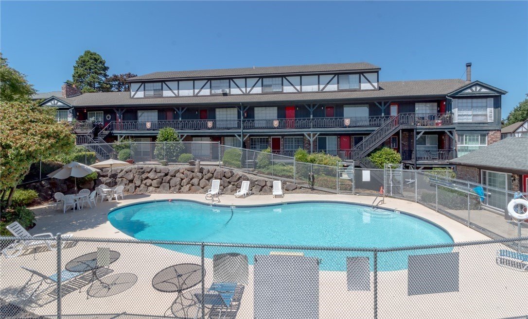 Apartment complex with a swimming pool, surrounded by a fence, and outdoor furniture including umbrellas and lounge chairs, under a clear blue sky.