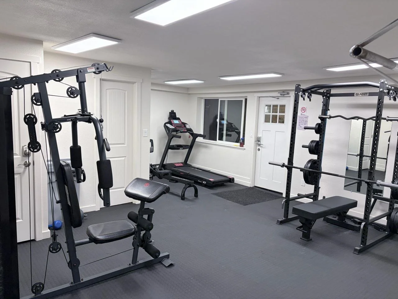 A small home gym with weightlifting equipment, a treadmill, and a window with a door next to it.