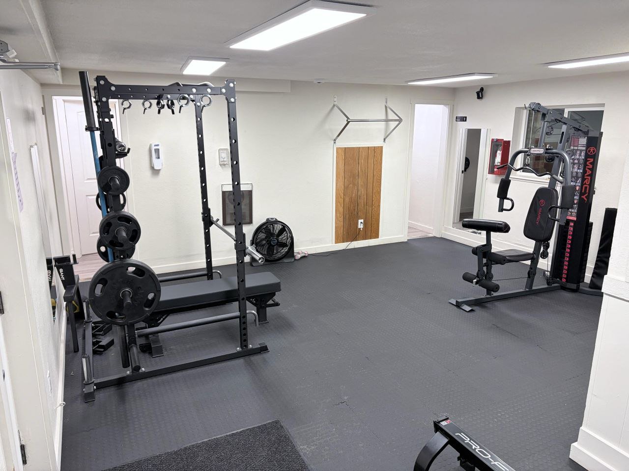 Home gym with weightlifting equipment, a bench press, a rowing machine, and a front-facing fan in a small room with white walls and ceiling lights.