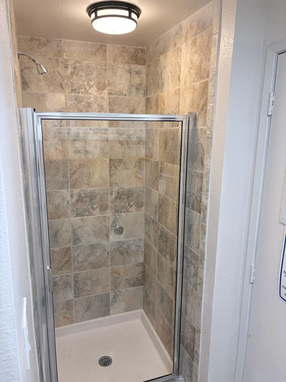 Shower with beige stone tile walls, a glass door, silver frame, and a white shower base, overhead ceiling light