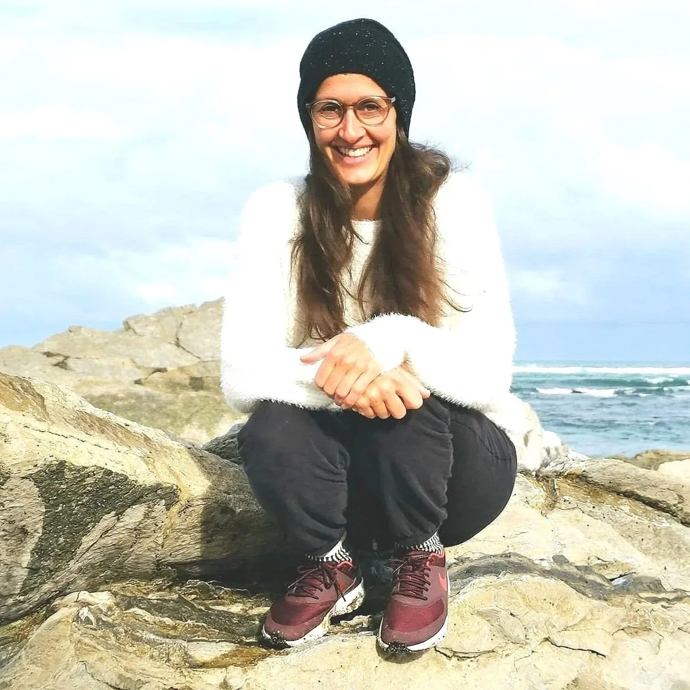 Sandra Jovanovic, Psychologische Beraterin am Meer, lächelnd auf einem Felsen. Blick in die Kamera.