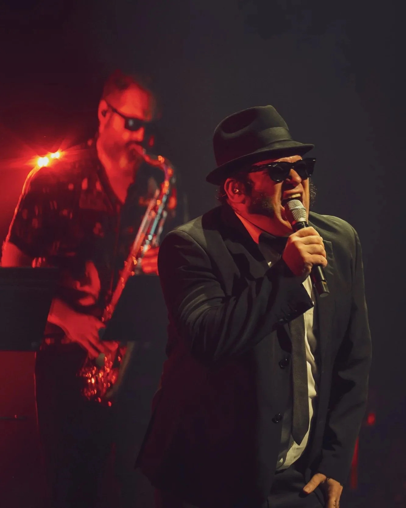 A man dressed in a black suit, black hat, and sunglasses singing into a microphone. A musician in the background is playing the saxophone, wearing glasses and a patterned shirt, with stage lighting creating a red glow.