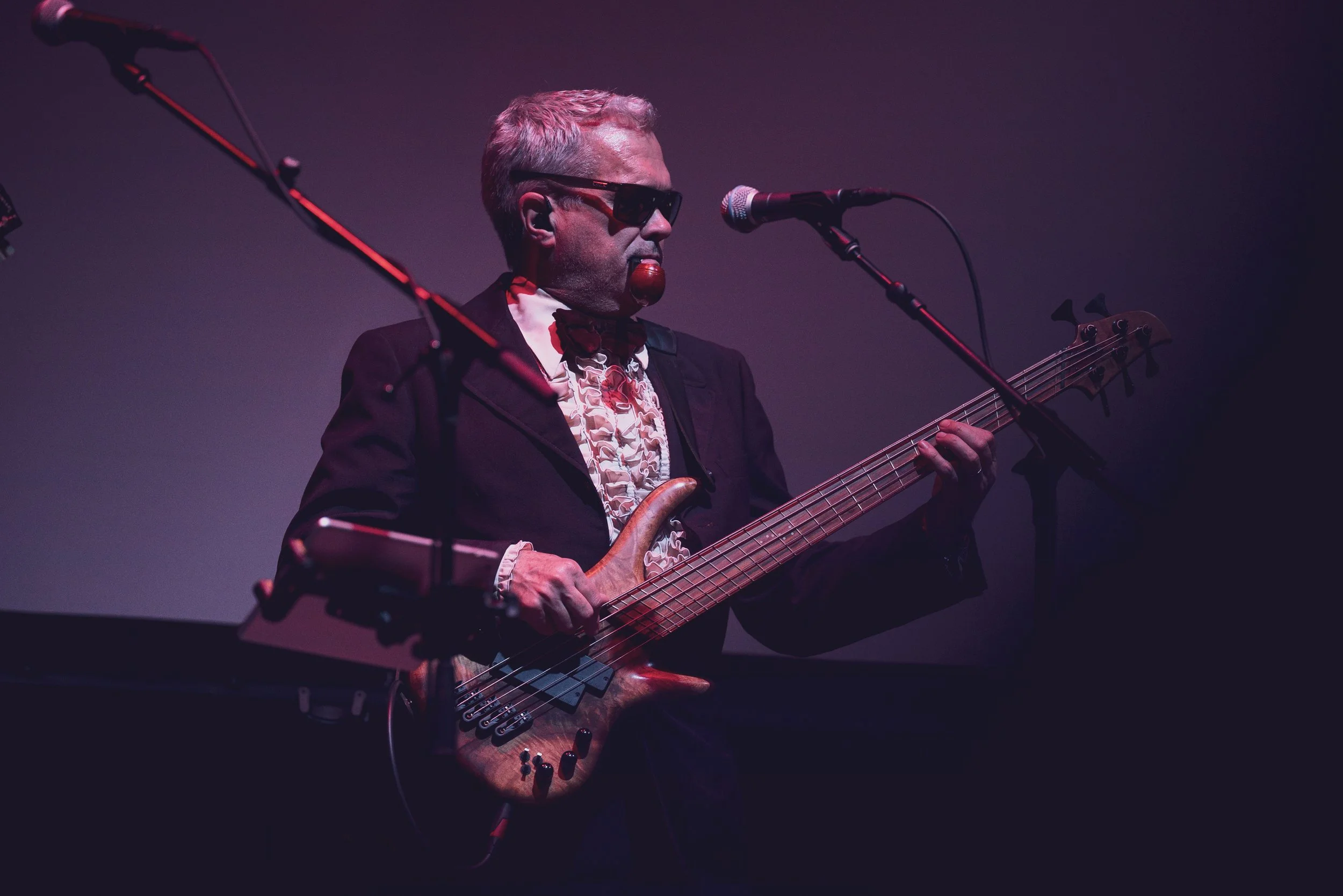 A man wearing dark sunglasses and a patterned shirt playing a wooden bass guitar on stage, with microphones positioned nearby under dramatic stage lighting.