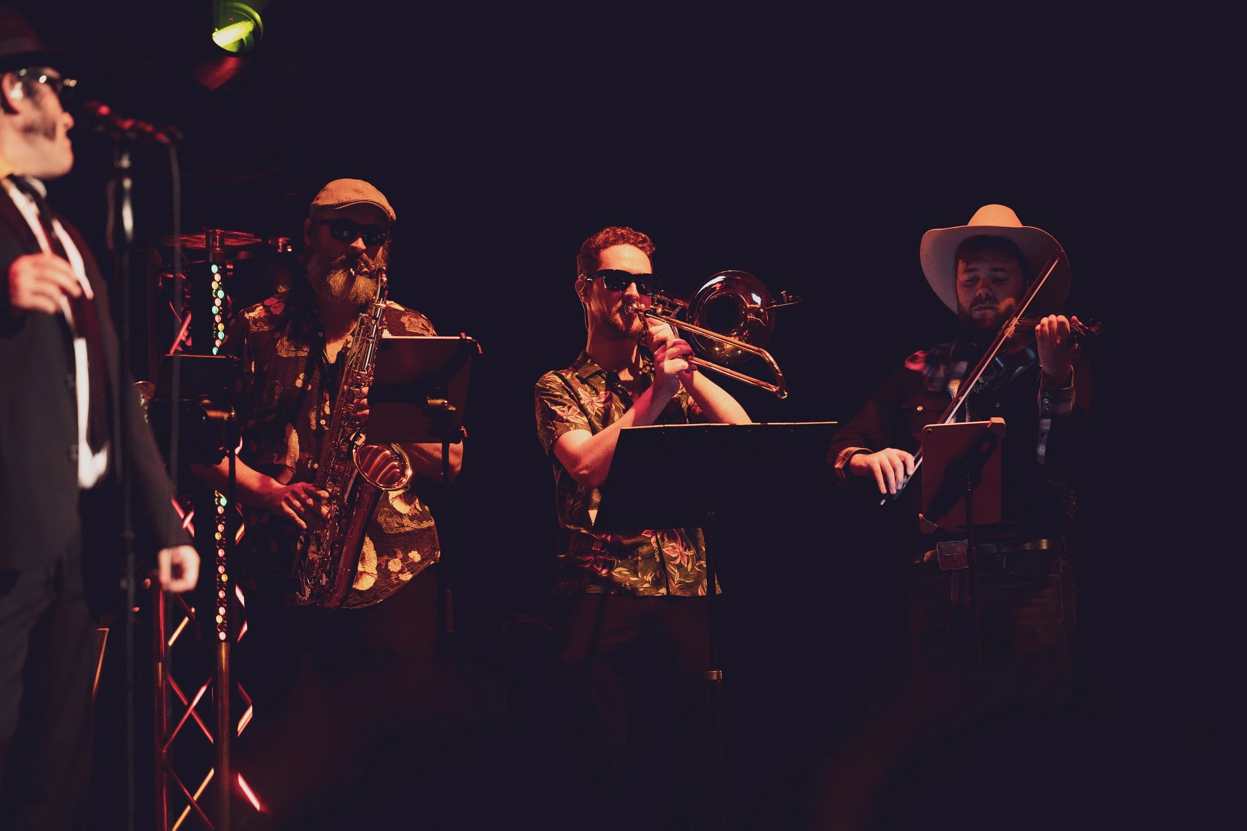 Musicians performing on stage, including a saxophonist, a trombonist, and a violinist, in a dark setting with stage lighting.