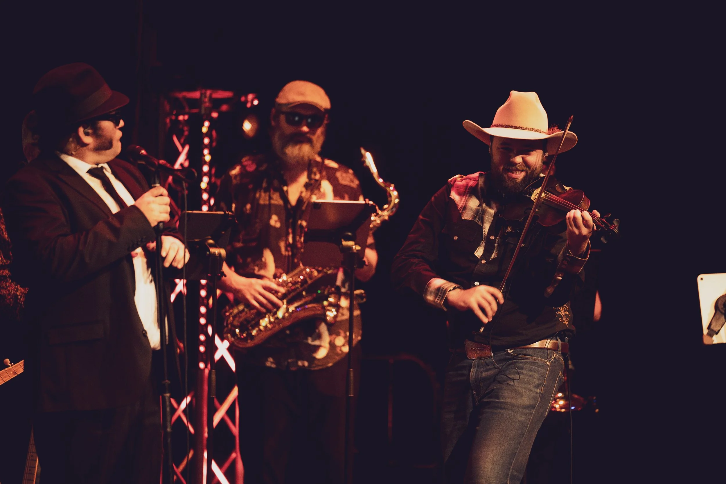 Three musicians playing instruments on stage in a dark setting: a man in a suit and hat singing into a microphone, a man with a saxophone wearing sunglasses and a hat, and a man with a violin smiling and playing with a hat and plaid shirt.