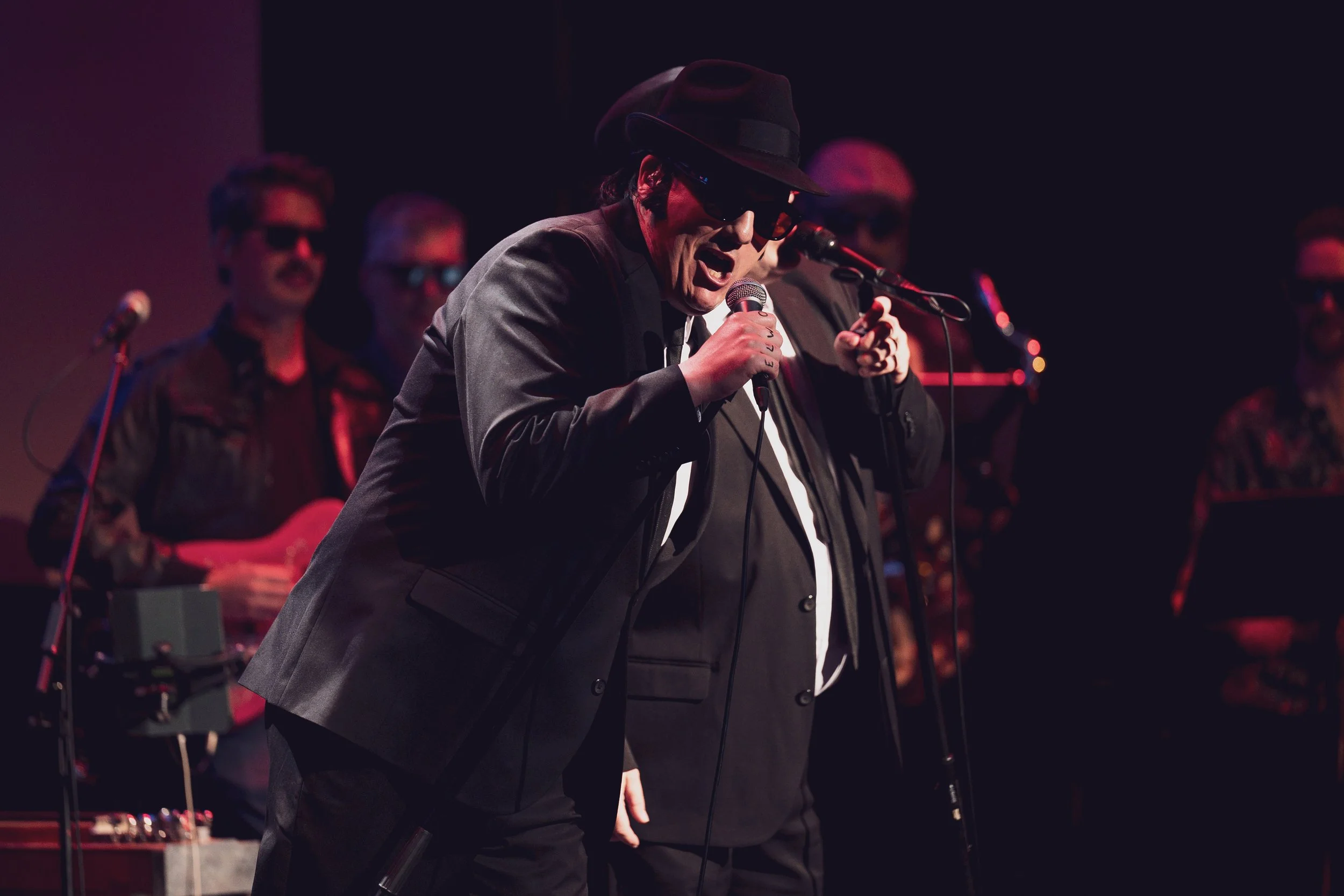 Man wearing black hat and sunglasses singing into a microphone on stage, with several backup performers in the background.