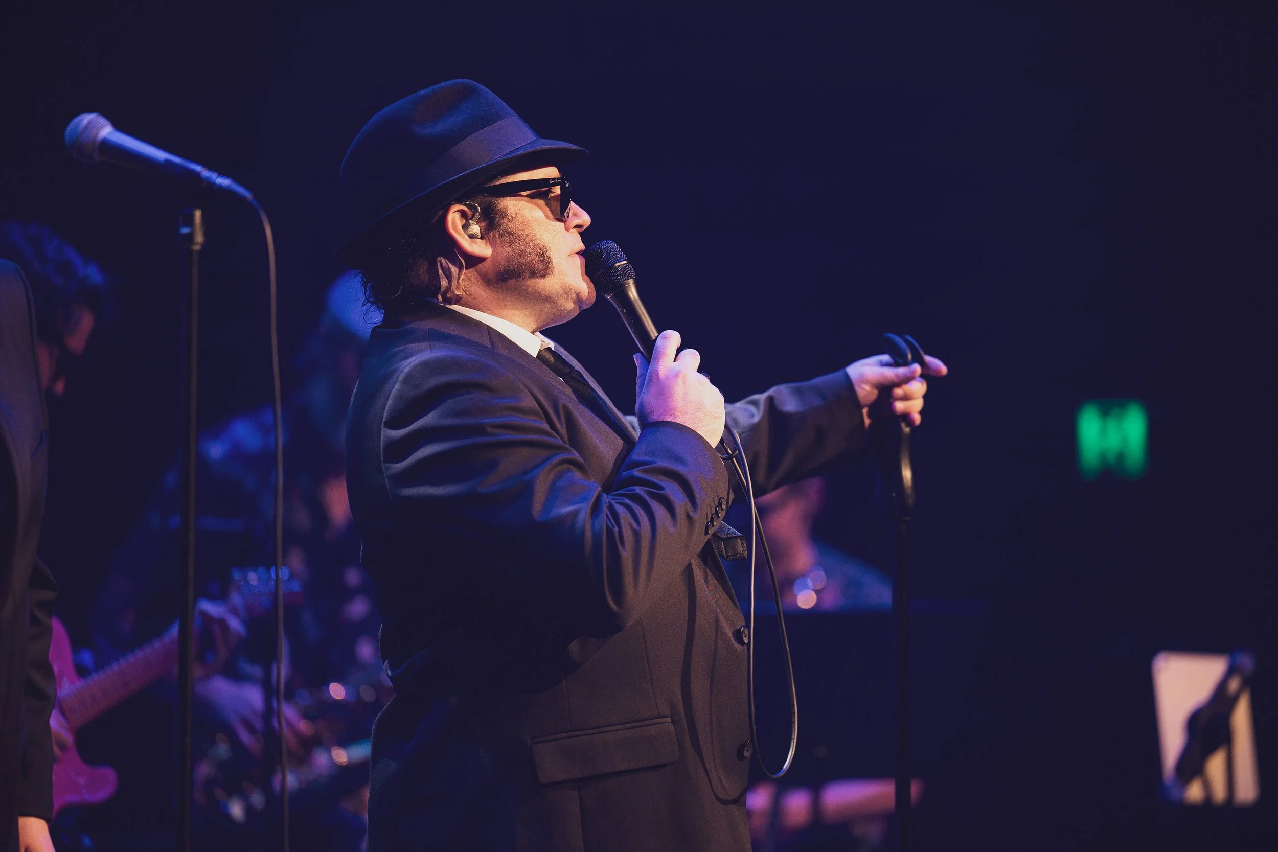 A man wearing a black hat, sunglasses, and a suit is singing into a microphone on stage during a performance.