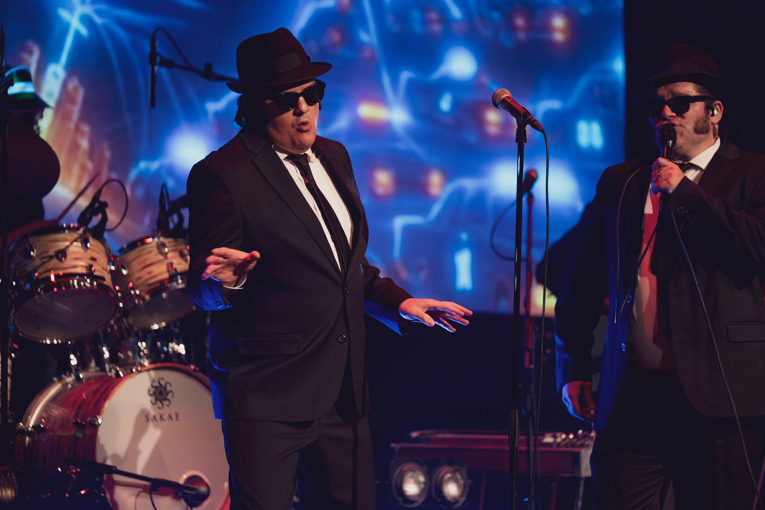 Two men in suits and hats performing on stage. They are wearing sunglasses and singing into microphones. Behind them, there are drums and a colorful, abstract digital background with blue and orange hues.