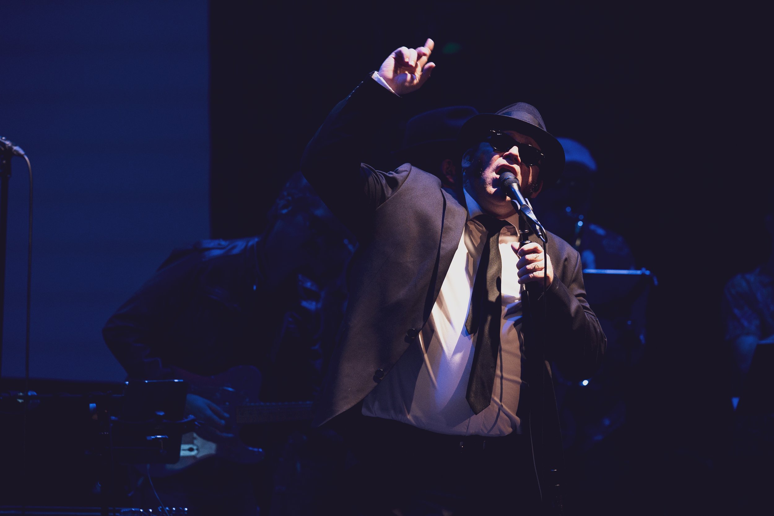 A man wearing a black fedora, sunglasses, and a dark suit with a white shirt and tie, singing into a microphone on stage with a dark background.