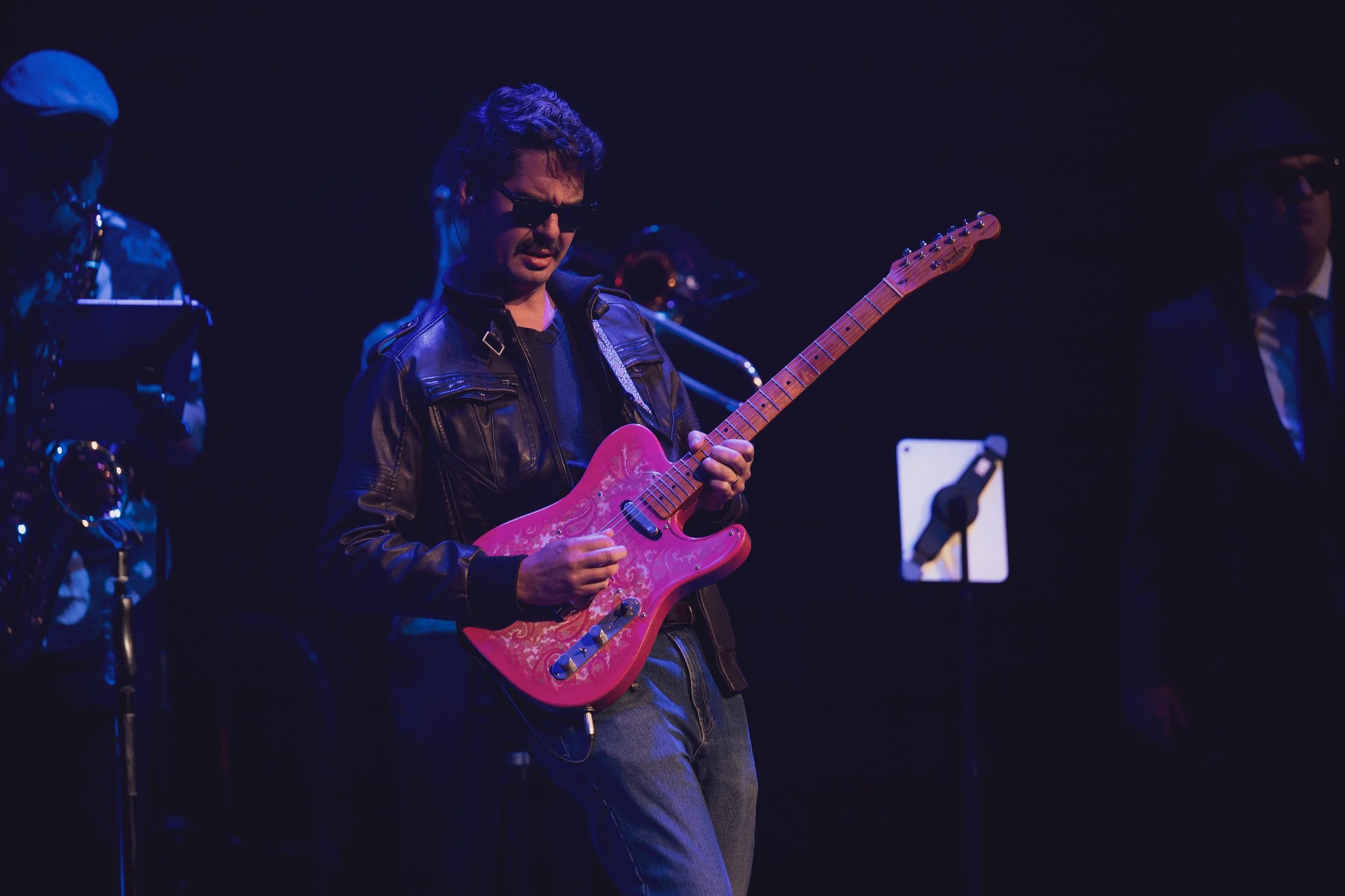 A man wearing sunglasses and a leather jacket playing a pink electric guitar on stage, with dark background and other band members partially visible.