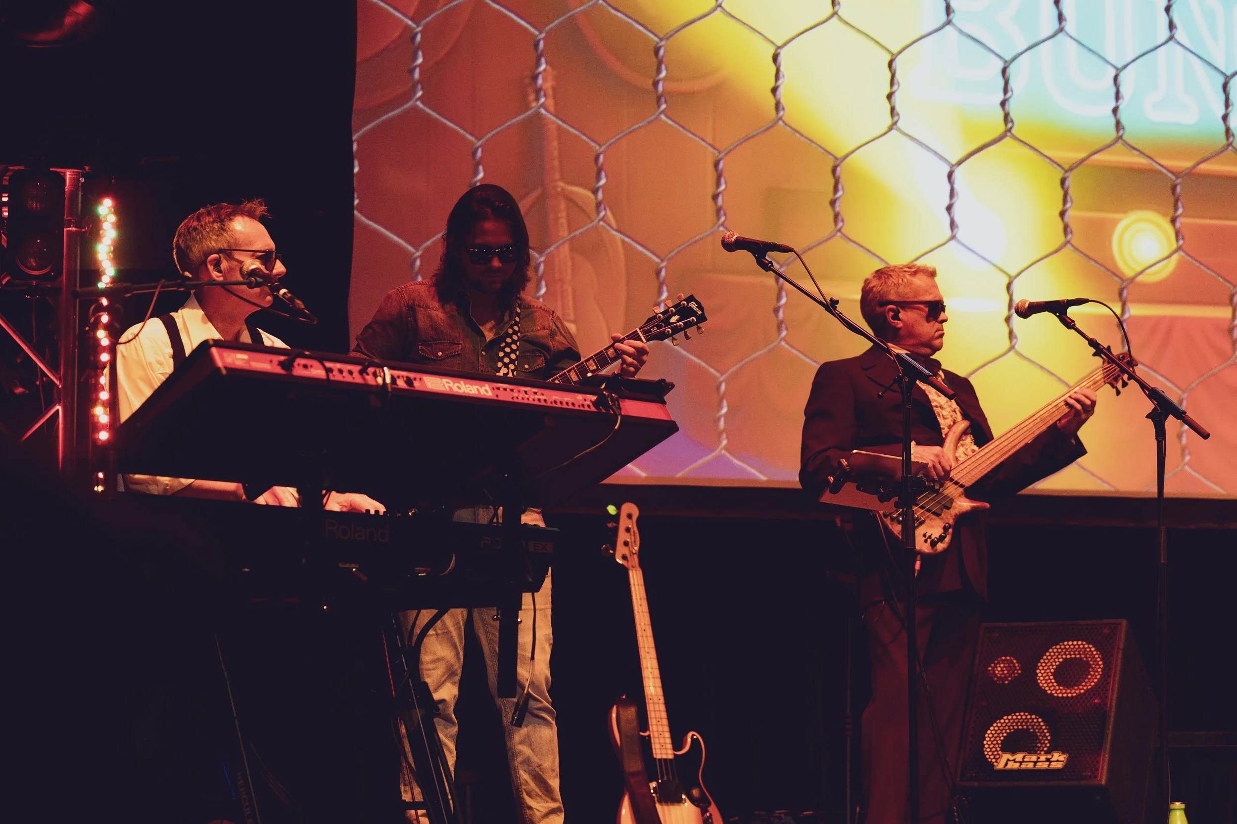 Three musicians perform on stage: one playing keyboard, another playing guitar, and the third playing bass. They wear sunglasses, and the background features a colorful, illuminated projection with a chain-link pattern.