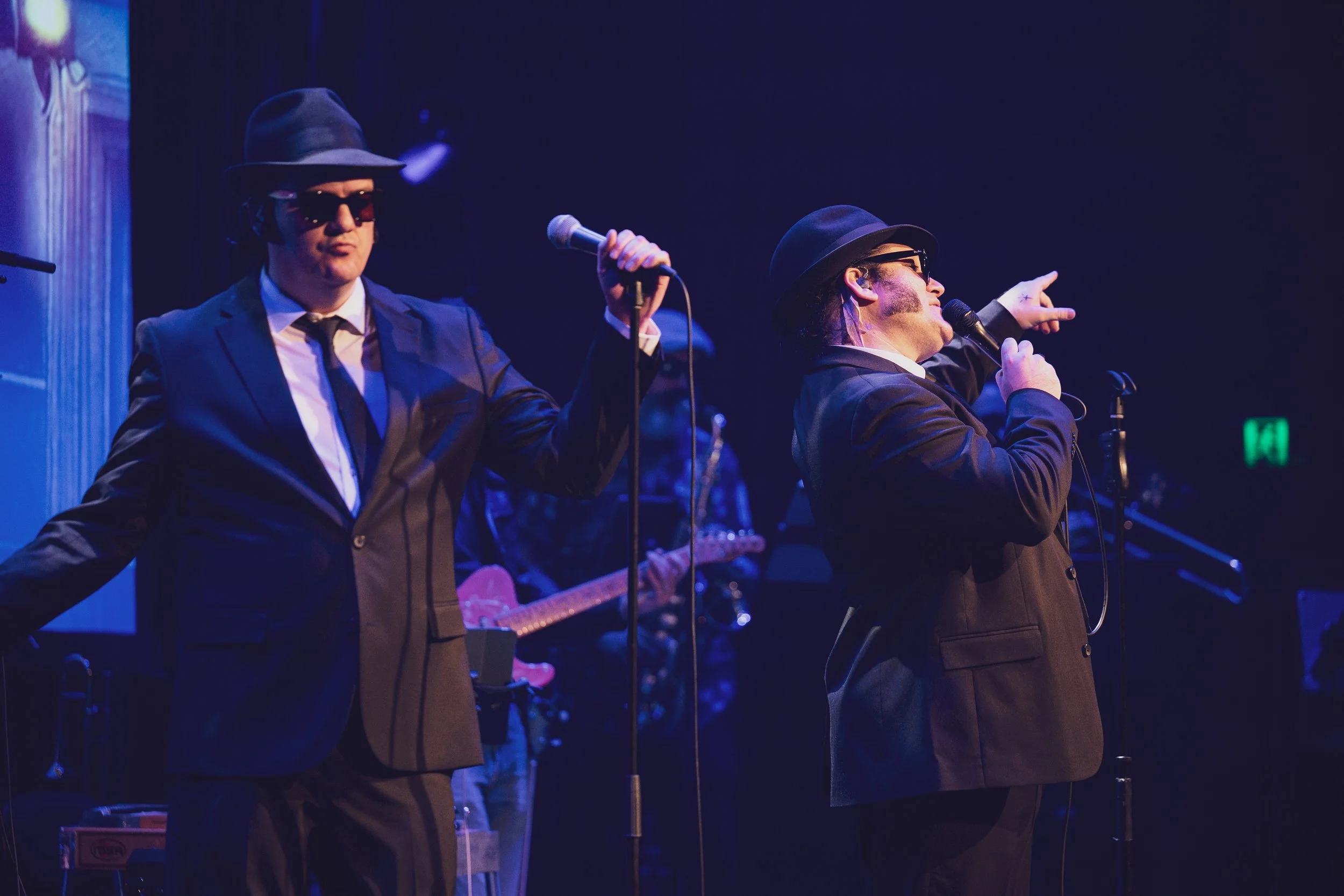 Two men wearing black suits, black hats, and sunglasses performing on stage with a microphone. One is pointing to the right, and there is a musician in the background playing a guitar.