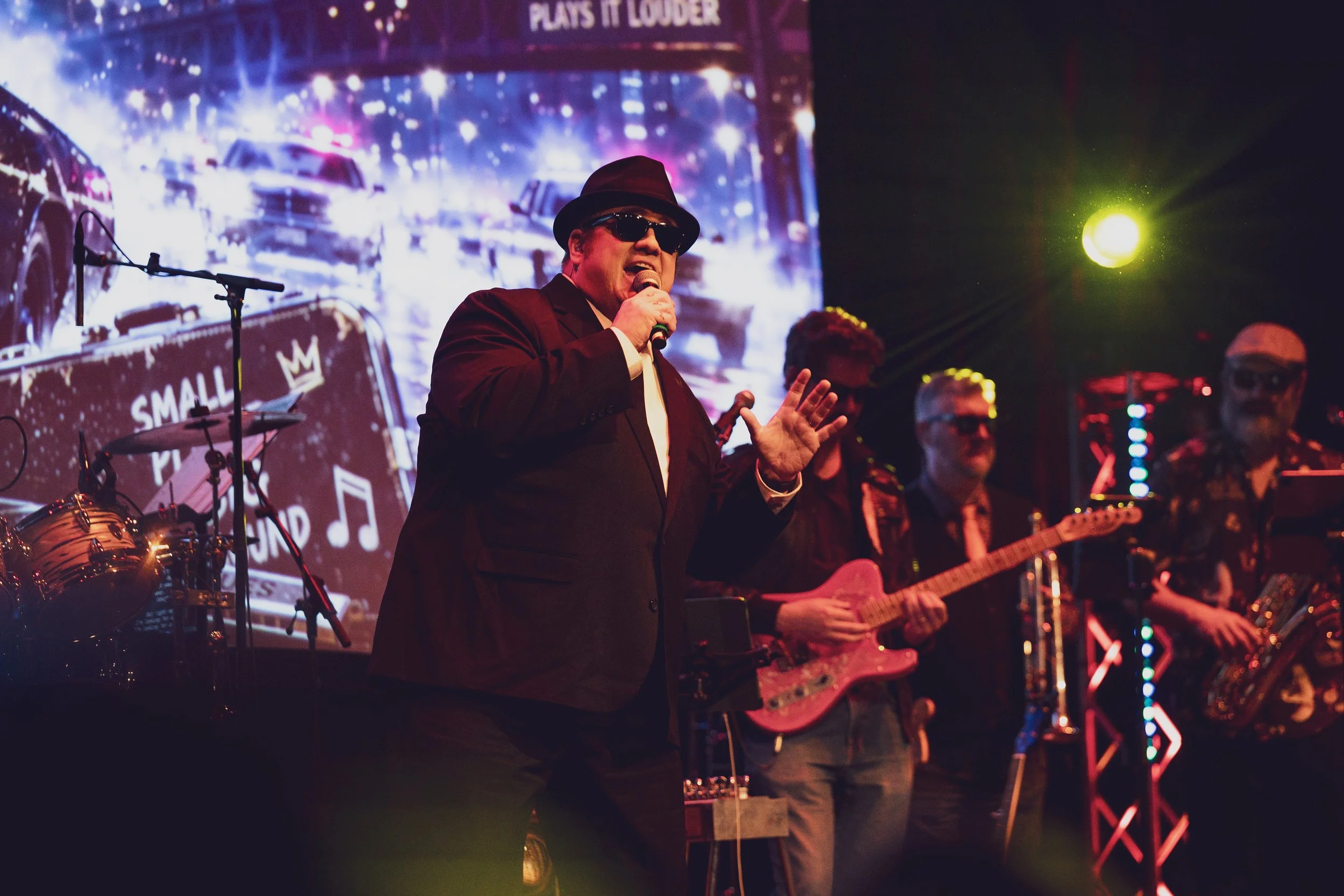 A man wearing sunglasses and a hat singing into a microphone on stage, with a band and bright colorful lights in the background.
