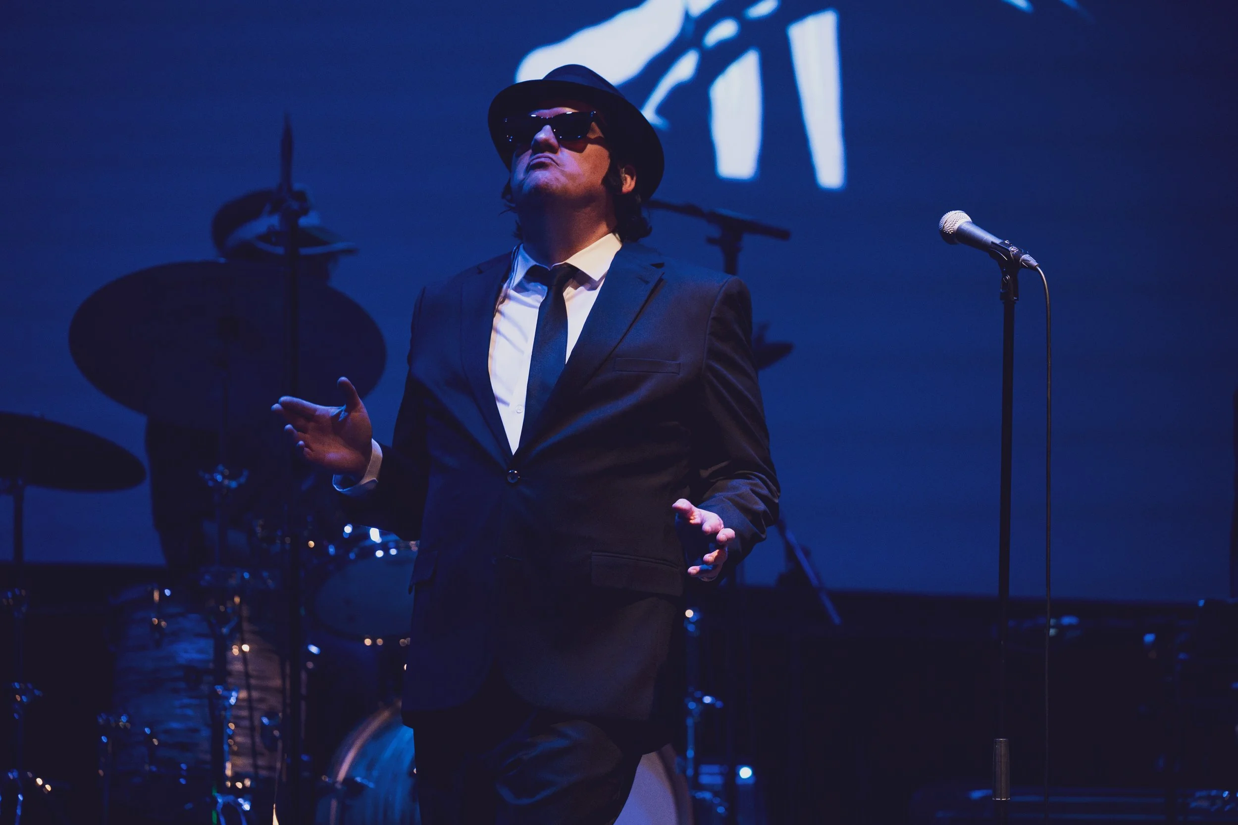 A man dressed in a black suit, white shirt, black tie, wearing sunglasses and a black fedora, standing on stage with a microphone and musical instruments in the background.
