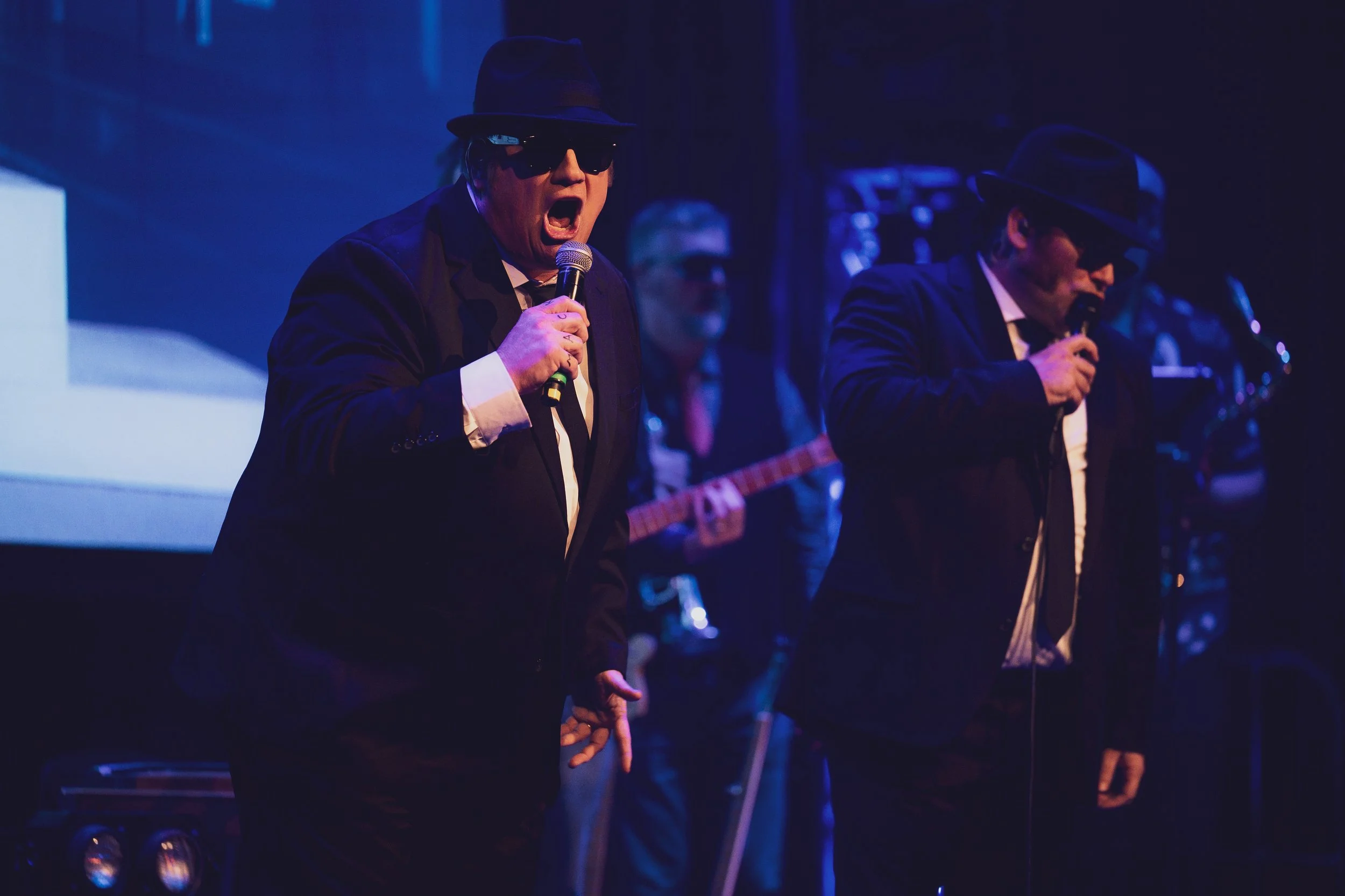 Man in a suit singing into a microphone with others in similar attire holding microphones on stage, all wearing dark suits, hats, and sunglasses, with blue lighting and a dark background.