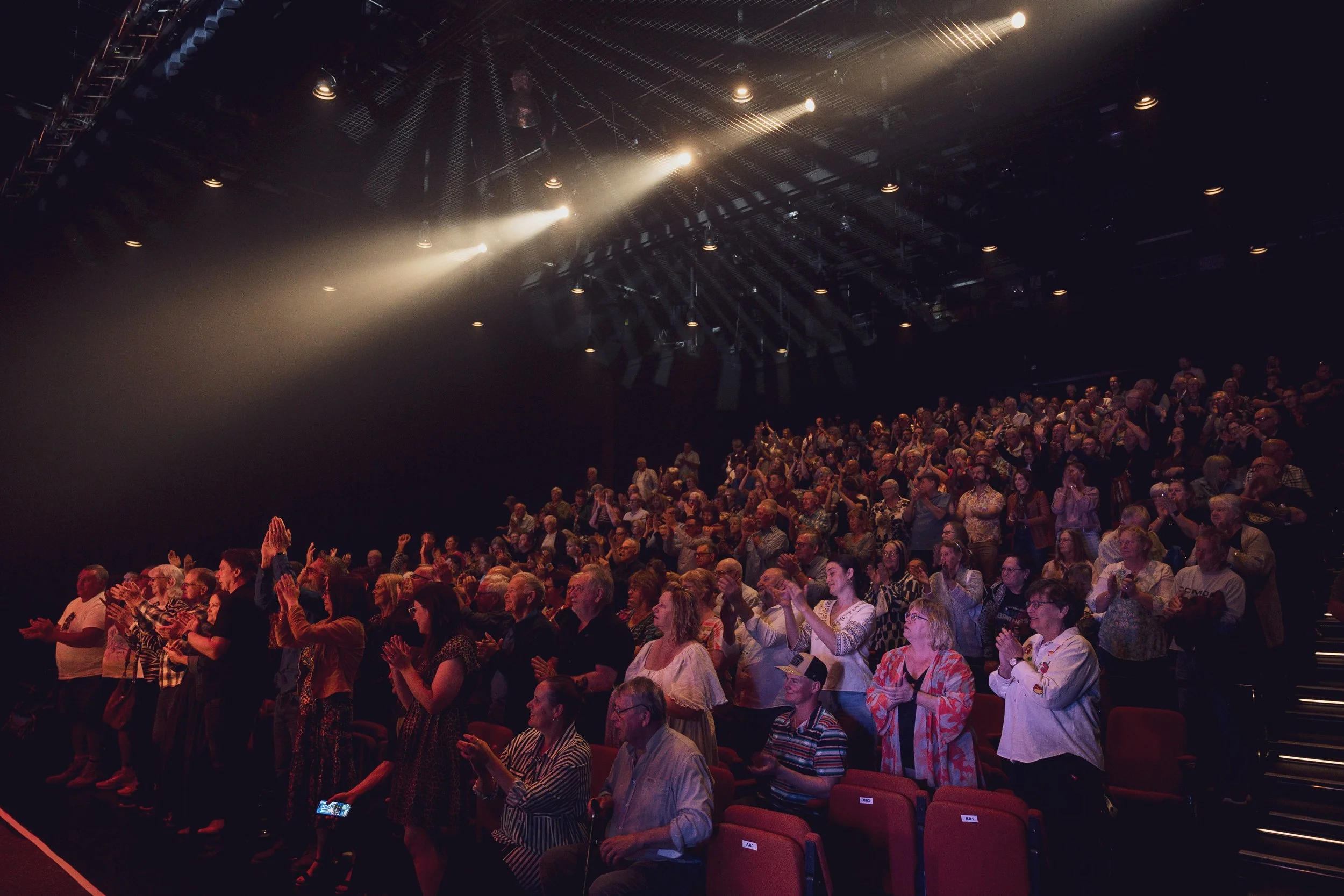 A large audience sitting in an auditorium with some people standing, clapping, and taking photos. There are bright stage lights shining down from the ceiling, which is fitted with a grid of stage lighting and rigging.
