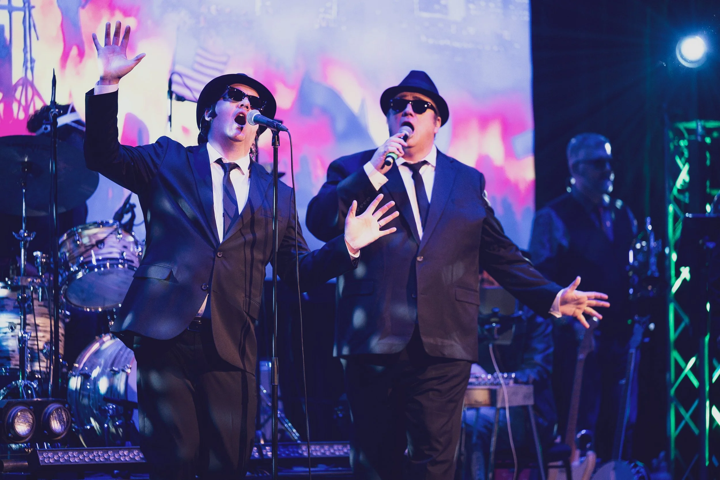 Two male performers in black suits, white shirts, black ties, and sunglasses singing on stage with microphones. They are accompanied by a drummer and other band members in the background, with colorful stage lighting and a vibrant backdrop.