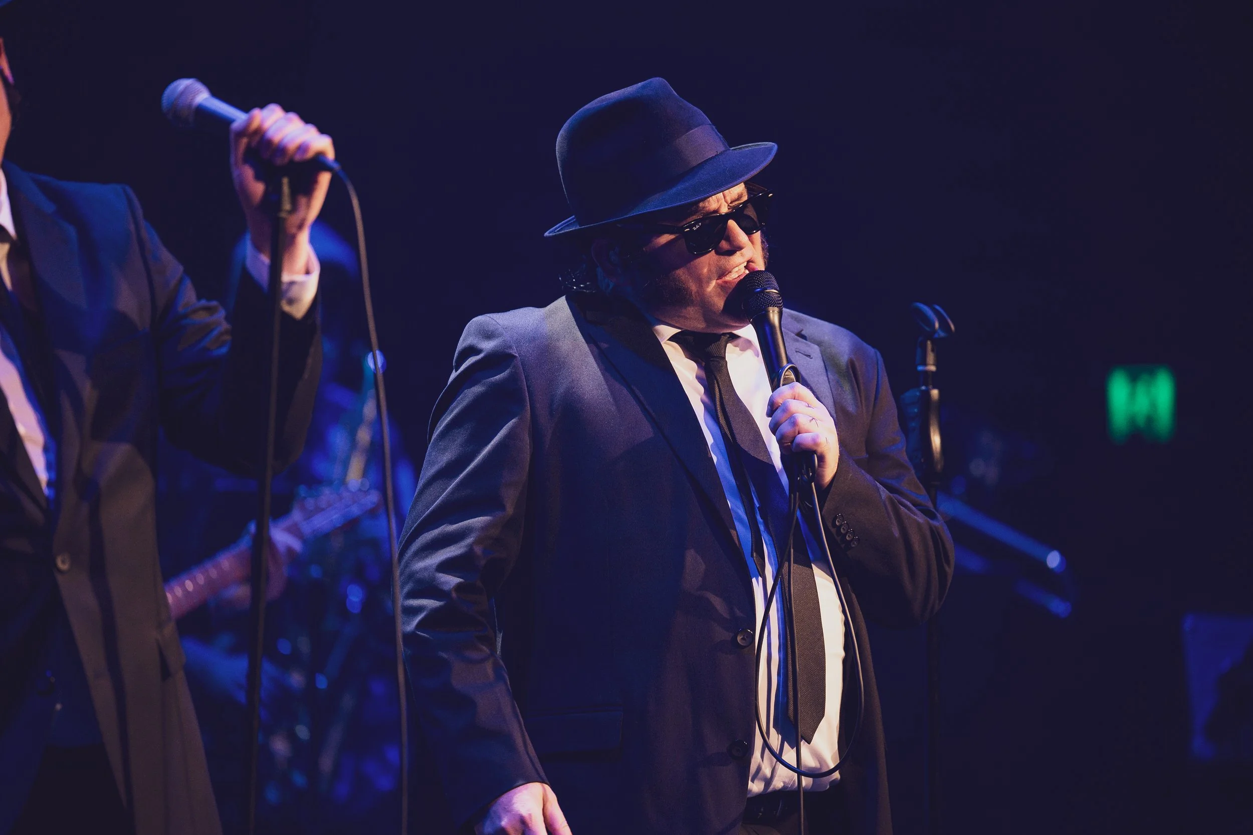 A man wearing a black suit, white shirt, black tie, black sunglasses, and a black fedora hat singing into a microphone on stage.