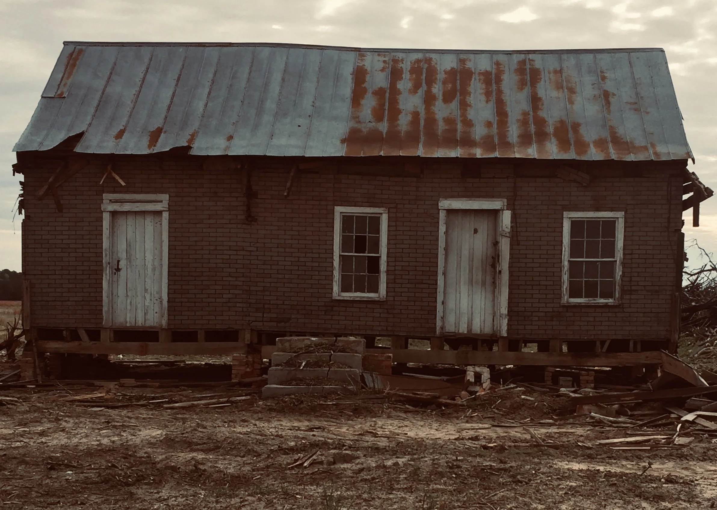 An old brick house with a rusted metal roof, window, and door, elevated on wooden stilts with debris and dirt in front.