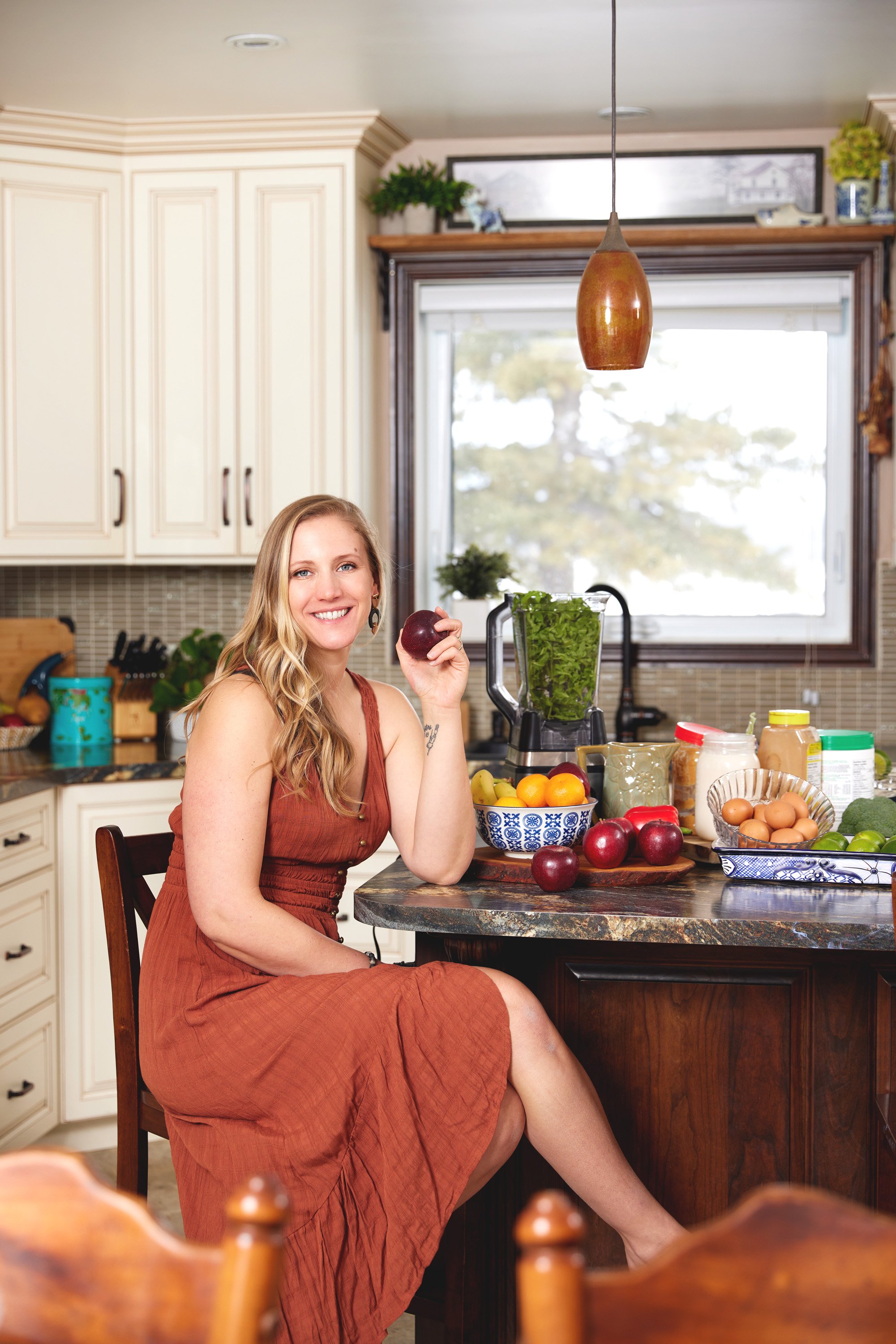 A smiling woman sitting in a kitchen, holding an apple, with a variety of fruits, vegetables, and kitchen items on the counter.