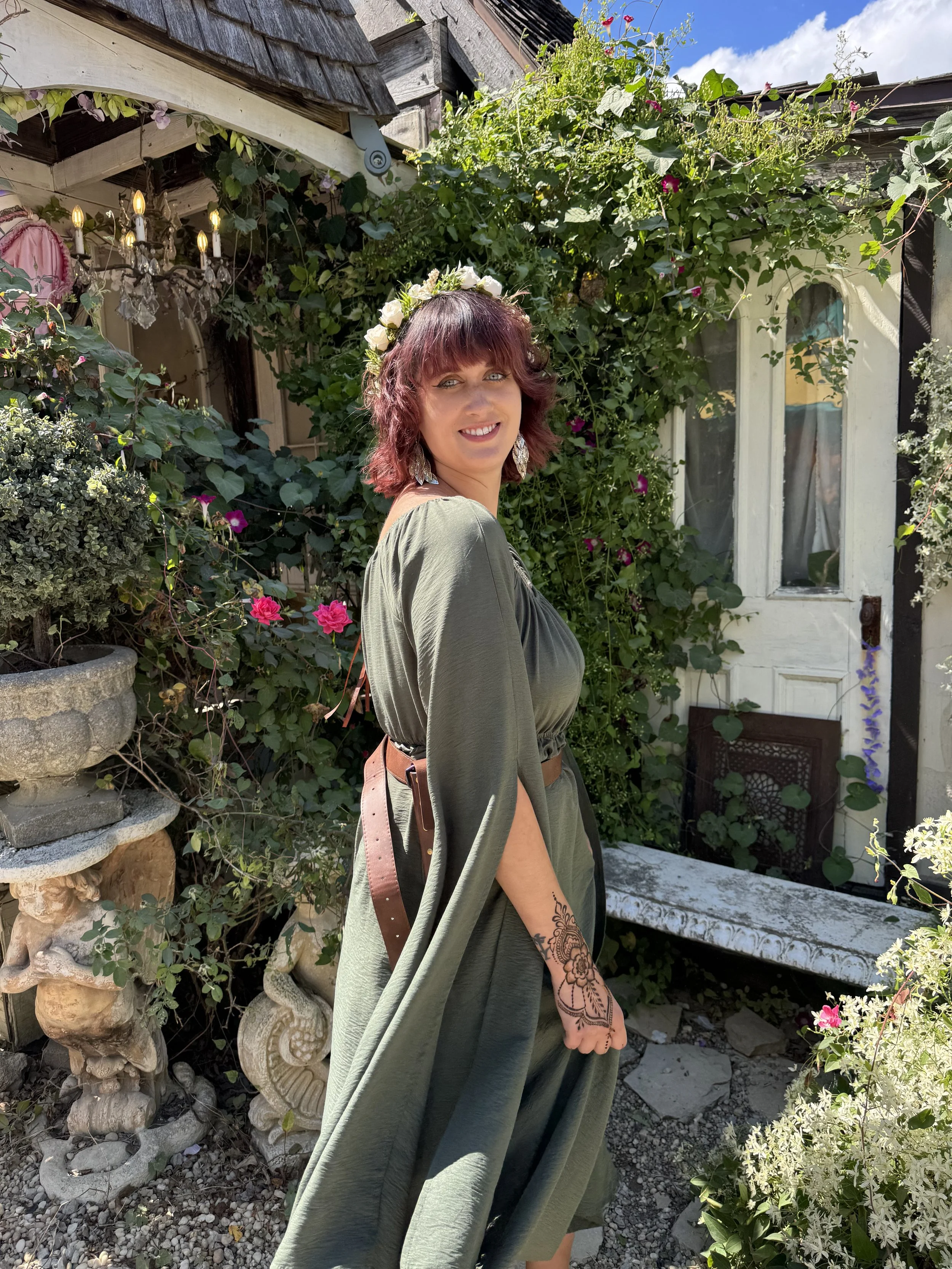 A woman with a flower crown and earrings standing in a garden with lush greenery and pink flowers, in front of a white door and a bench.