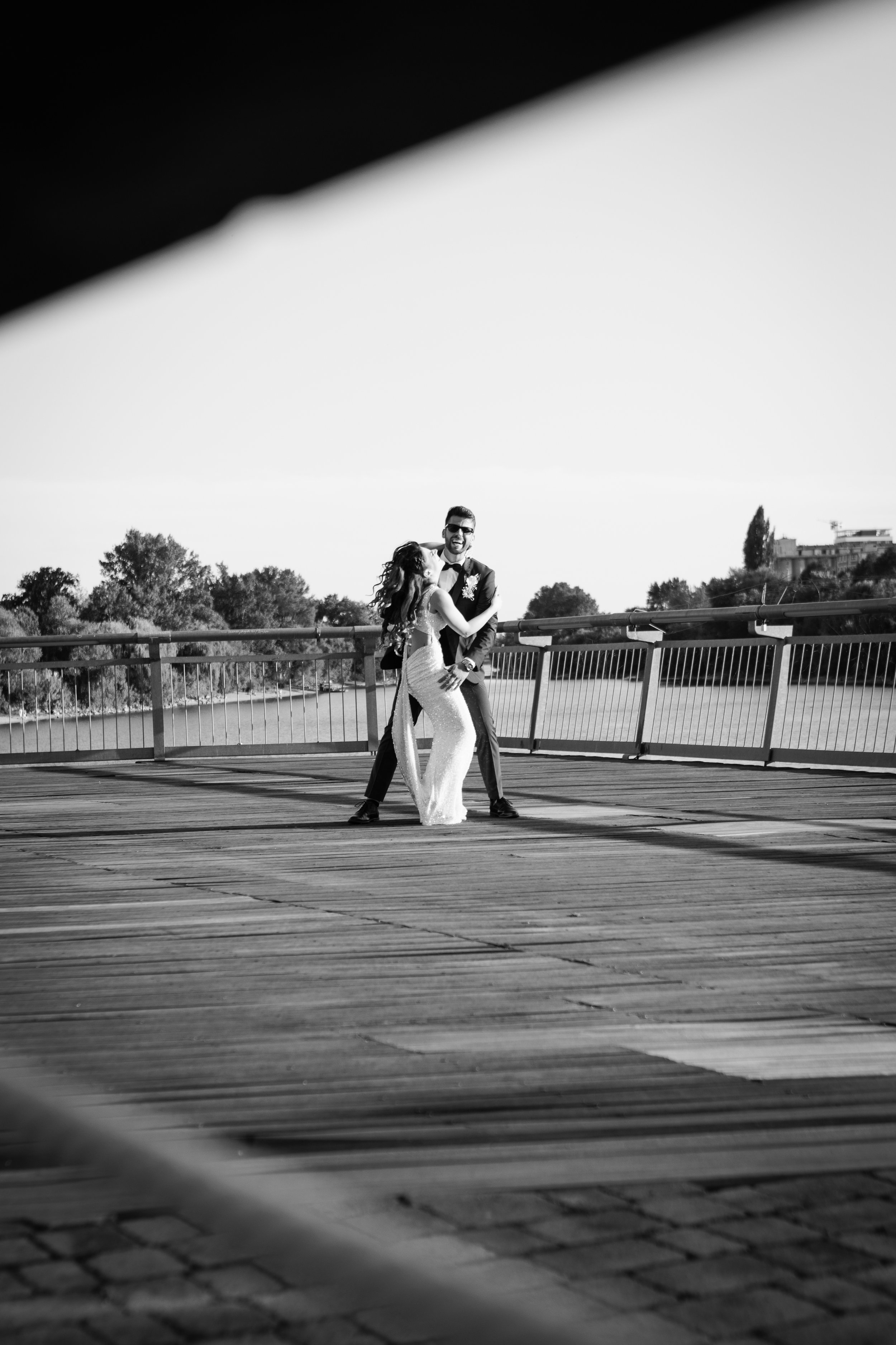 A black and white photo of a couple dancing on a wooden bridge, with trees and houses in the background.