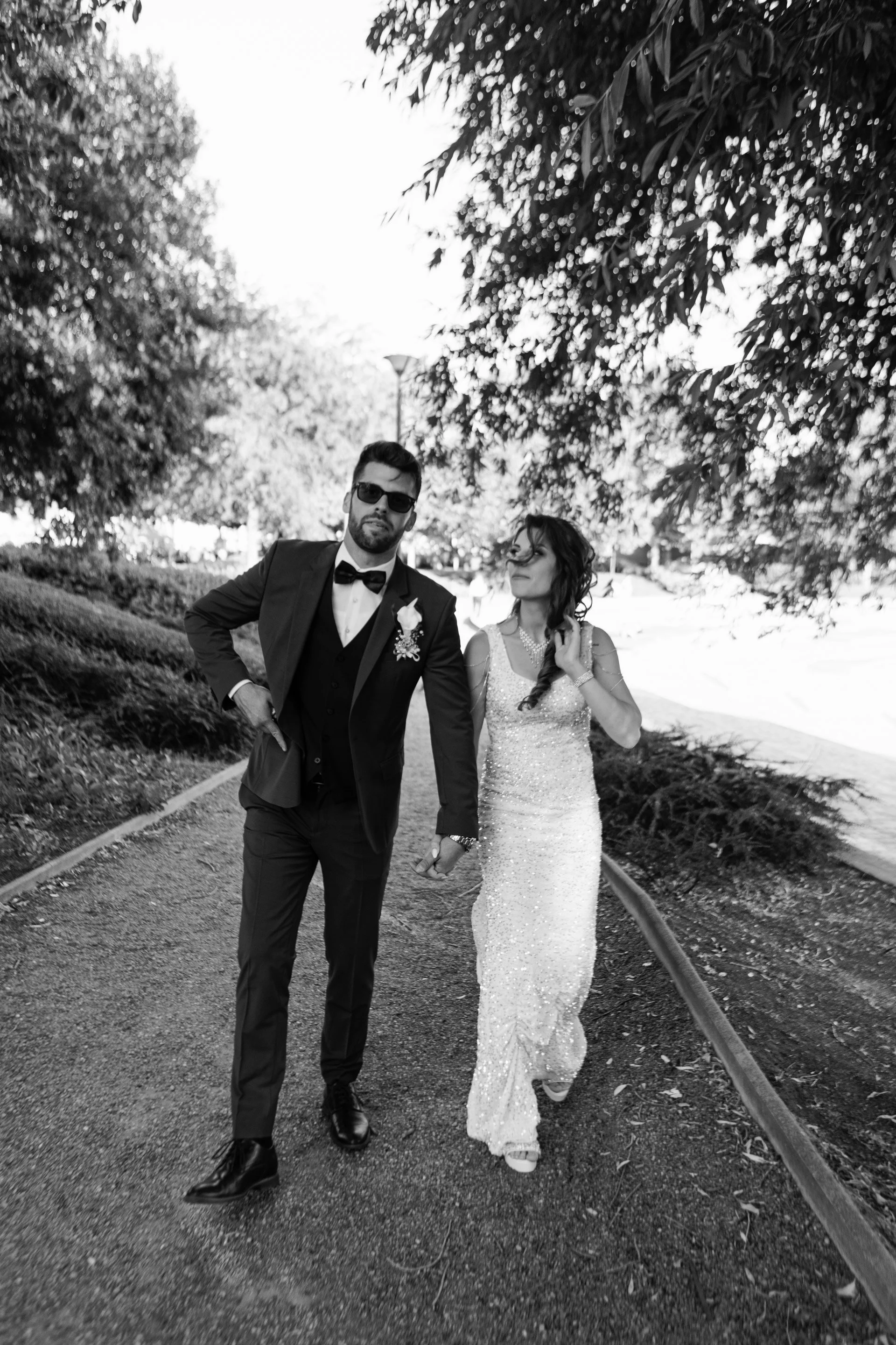 Black and white photo of a man and woman walking hand in hand on a park trail. The man is dressed in a tuxedo with sunglasses and the woman is wearing a long, sparkly gown and glasses.