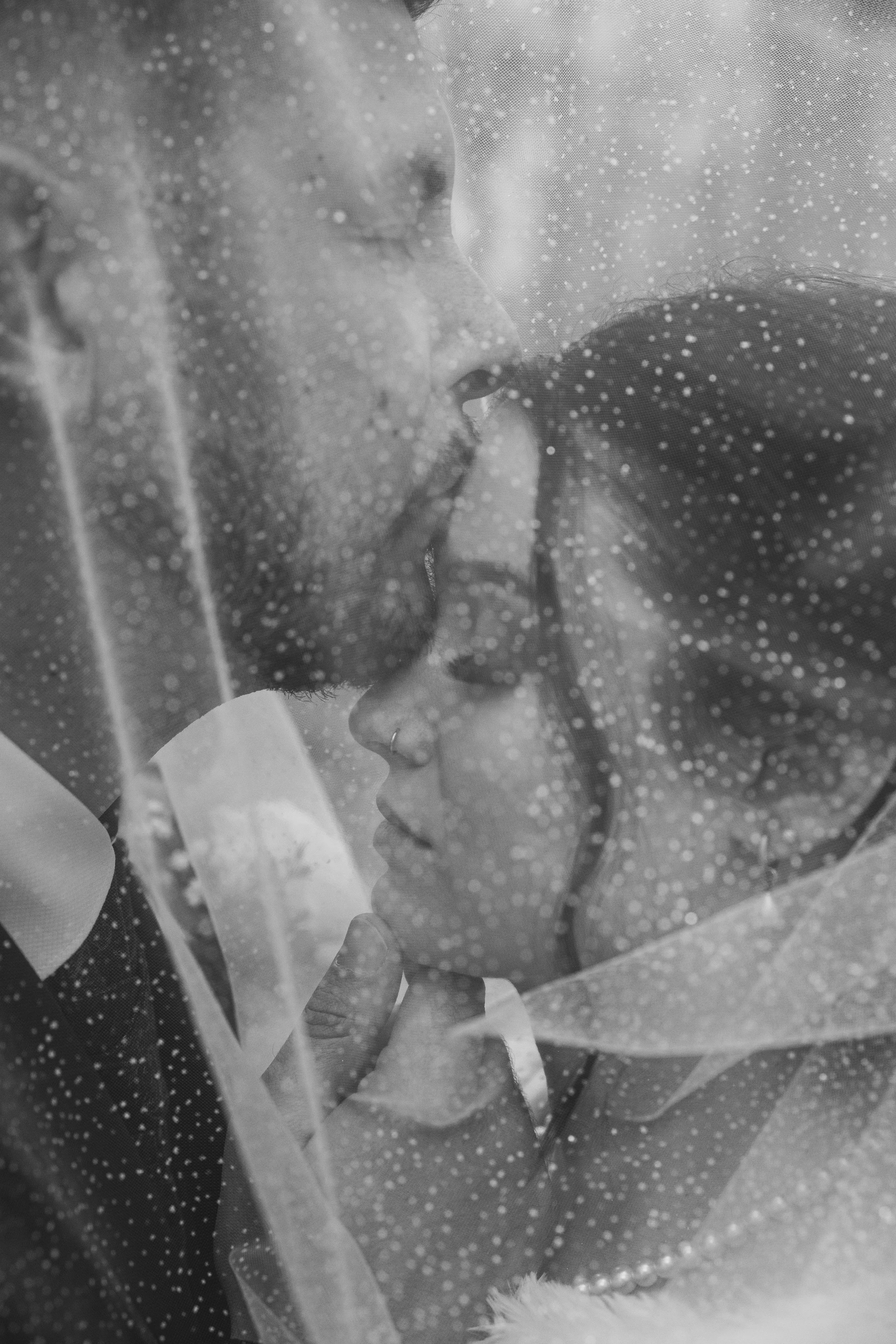 A black and white photograph of a couple kissing through a rain-covered window, with raindrops on the glass.