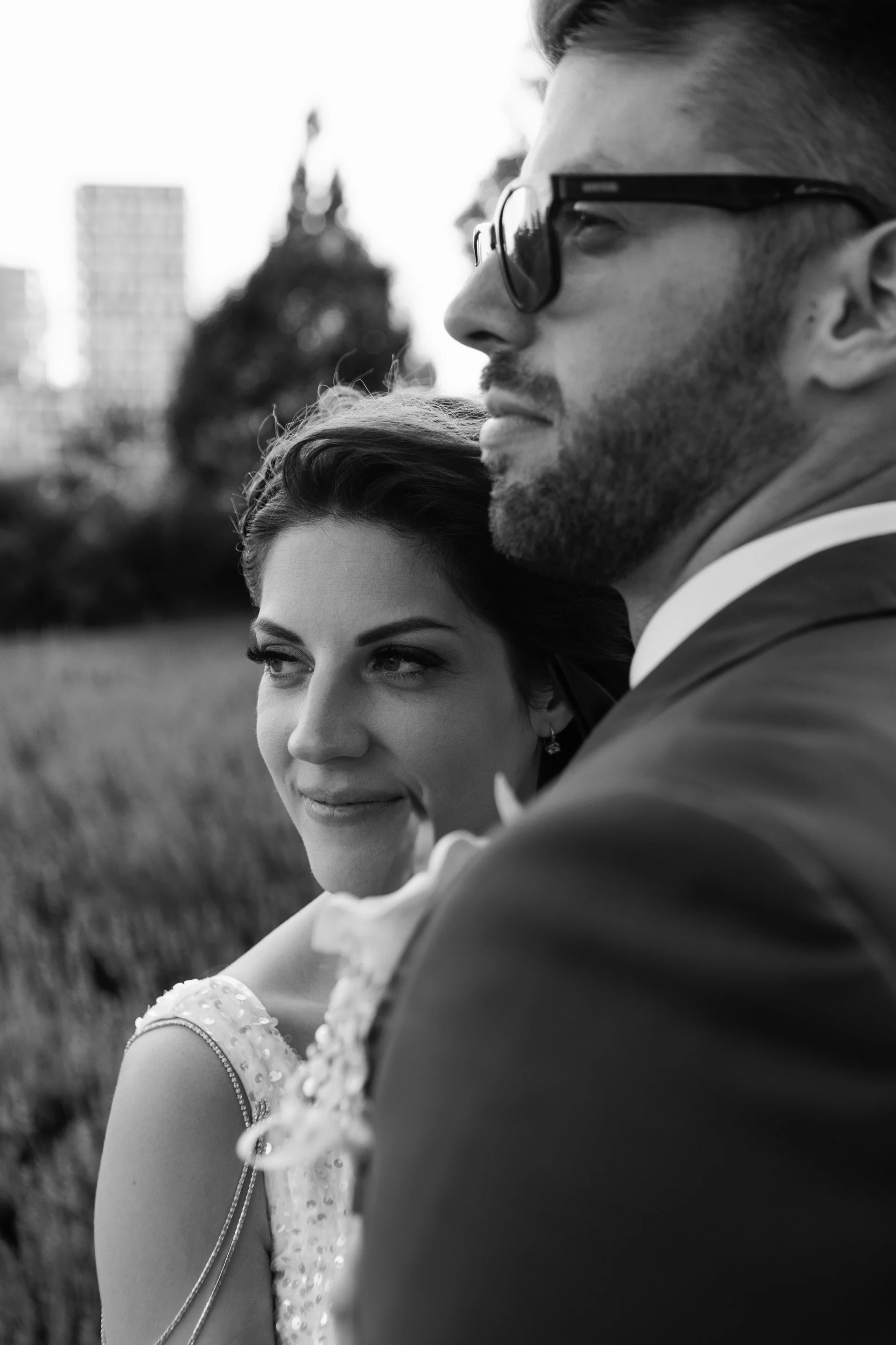 A black and white photo of a smiling woman and a man in a suit, standing outdoors with buildings in the background. The woman is looking at the man, who is wearing sunglasses.