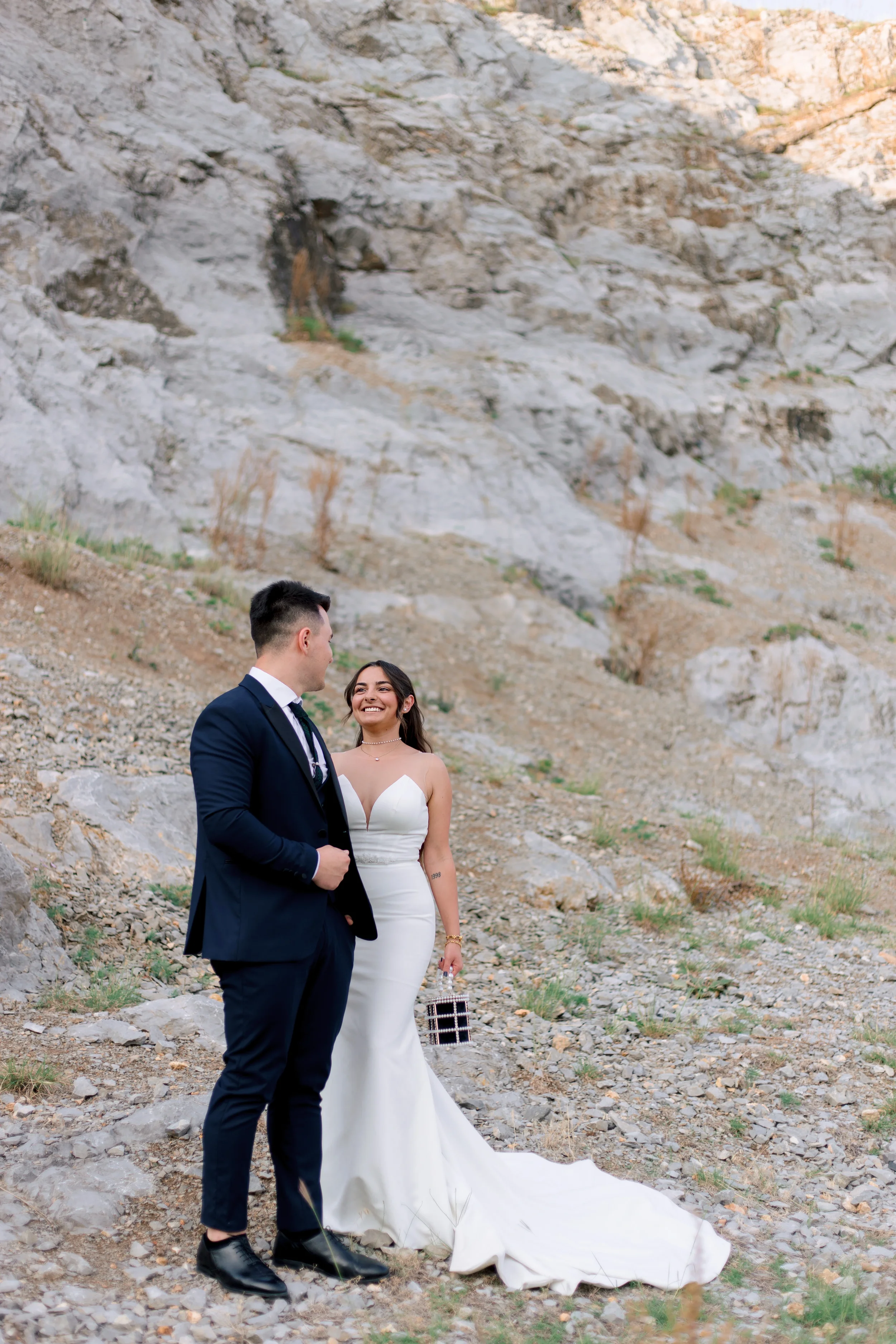 A bride in a white wedding dress and a groom in a dark suit standing outdoors near rocky terrain, smiling at each other.