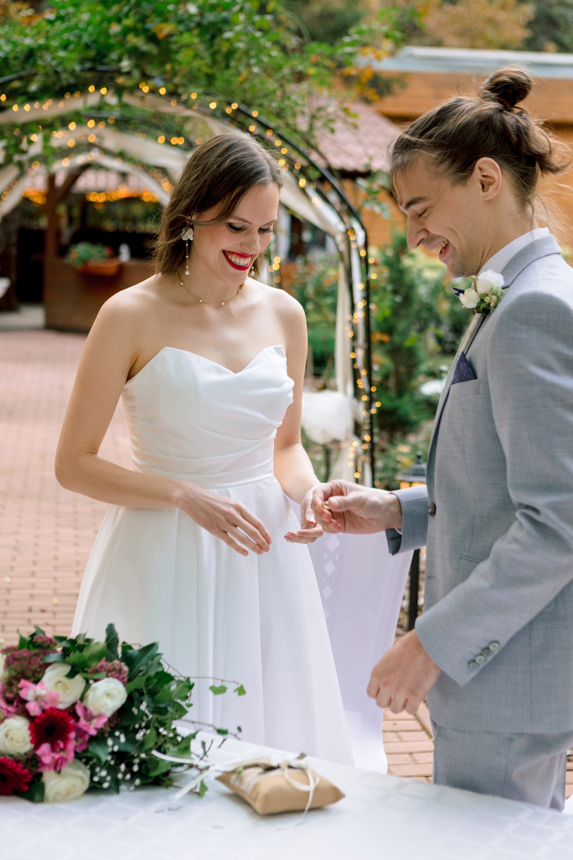 A bride and groom exchanging rings during their wedding ceremony outdoors, with flowers and a decorated arch in the background.