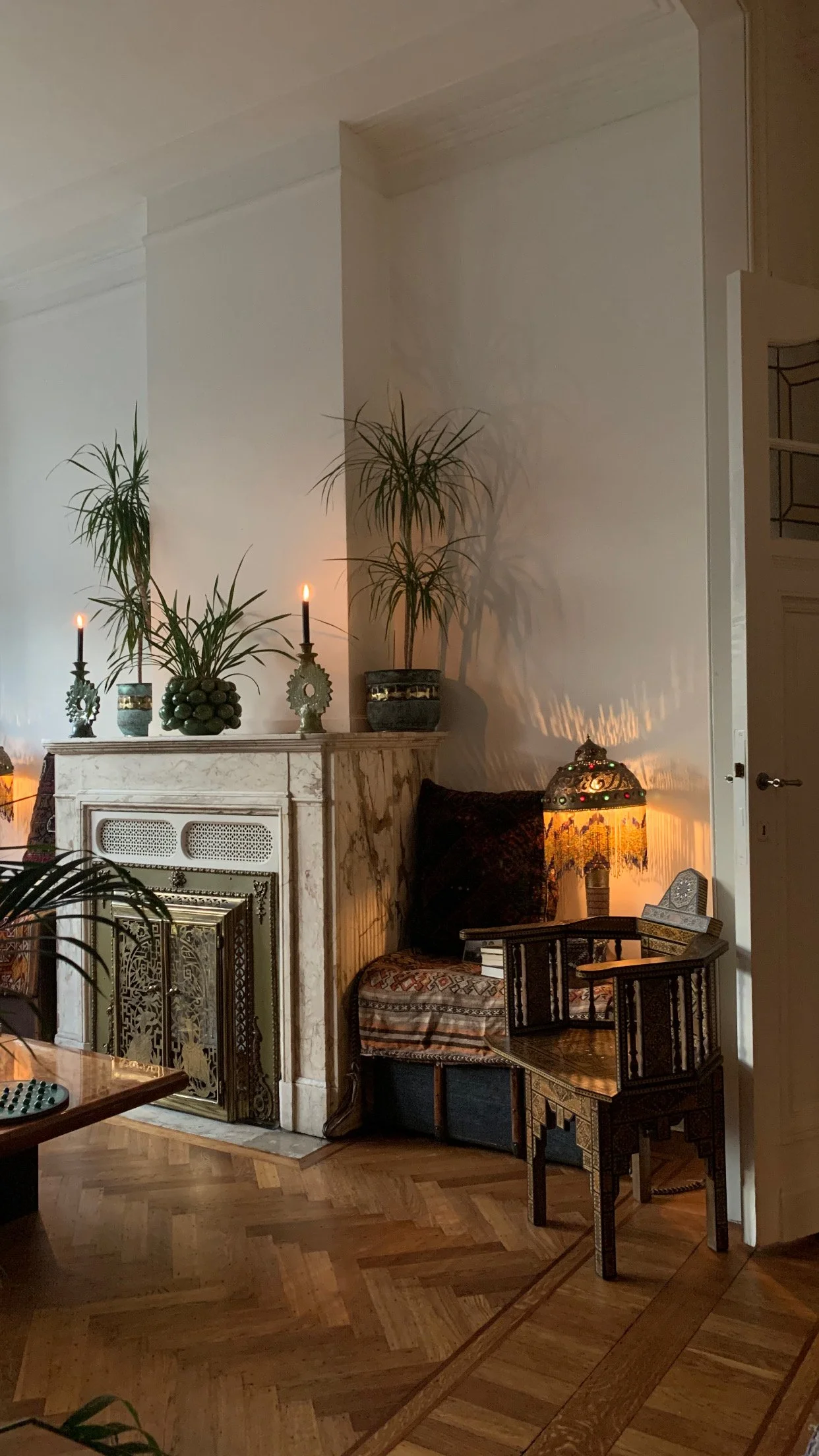 A cozy corner featuring a fireplace with a decorative gold screen, potted plants, candles, a vintage-style lamp with a stained-glass shade, a small wooden chair with detailed inlay and a cushion, and hardwood flooring with a geometric pattern.