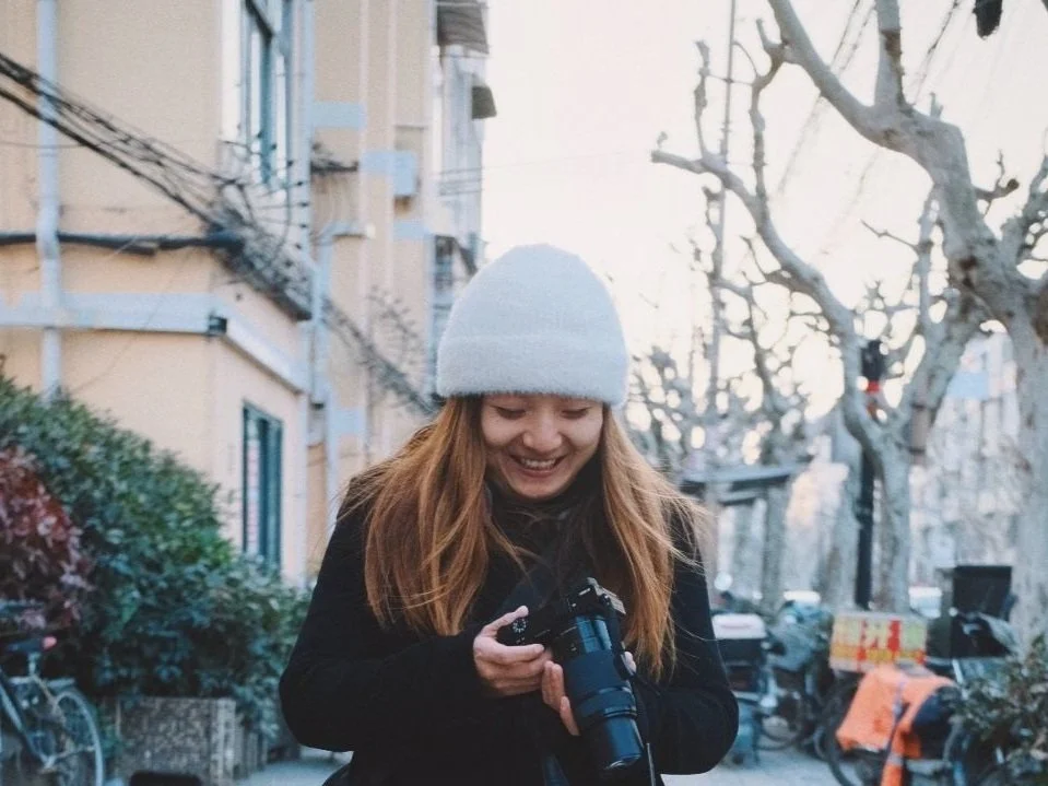 A young woman with long brown hair, wearing a white knit hat and black coat, smiling while looking at a camera she is holding outdoors in a city street with leafless trees and parked bicycles.