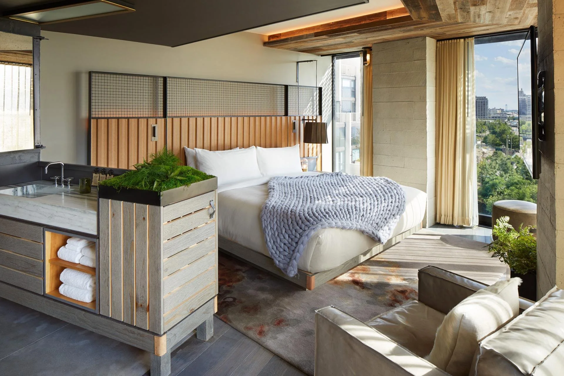 Modern hotel room with a large bed, cozy knitted blanket, wooden and metal design elements, greenery, and sunlight streaming through large windows with city views.