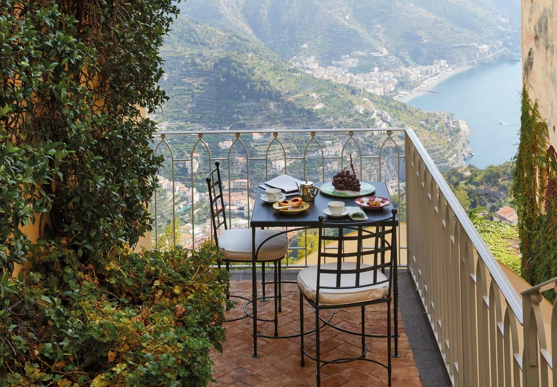 Balcony with table and chairs, breakfast setting, overlooking coastal town and sea.