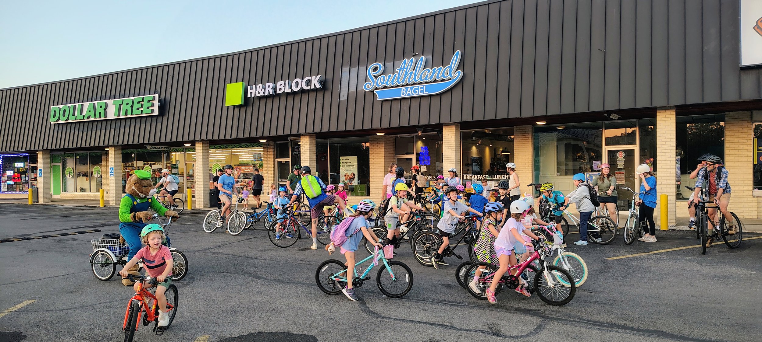 Students participating in the Picadome bike bus. There are a crowd of students and parents on bikes in front of Southland Bagel storefront. A wildcat mascot rides alongside on a tricycle.