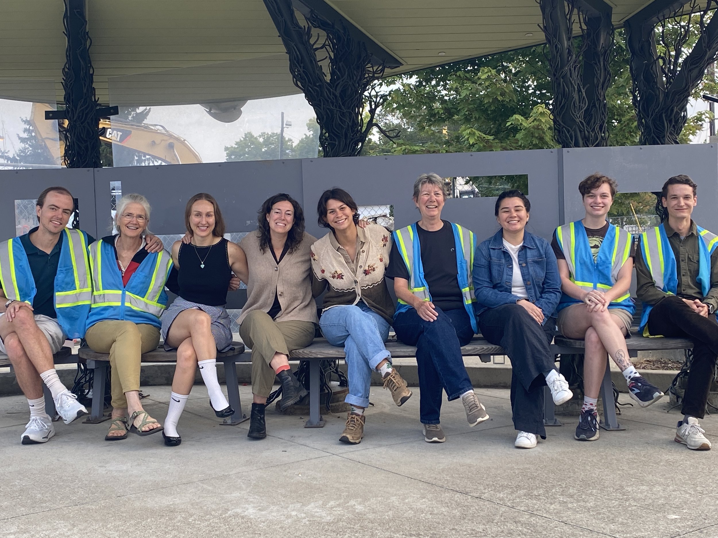 a group of nine people wearing safety vests, smiling and sitting at a bus stop.