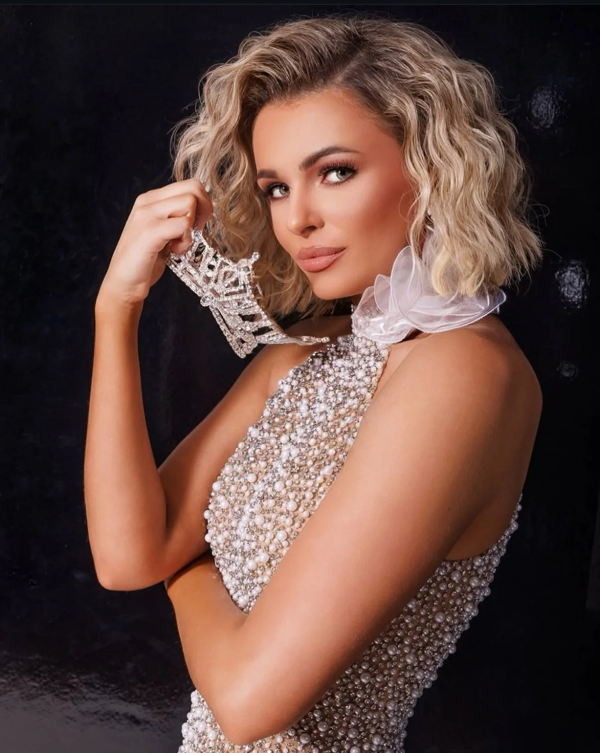 Woman with wavy blonde hair holding a large, ornate earring near her face, wearing a pearl and bead embellished dress with a sheer ruffle collar, against a black background. Miss America 
