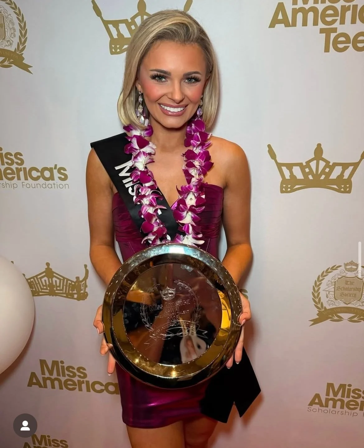 A woman in a purple dress holding a framed award, wearing a sash and a lei made of purple and white flowers, standing in front of a backdrop with 'Miss America' logos.