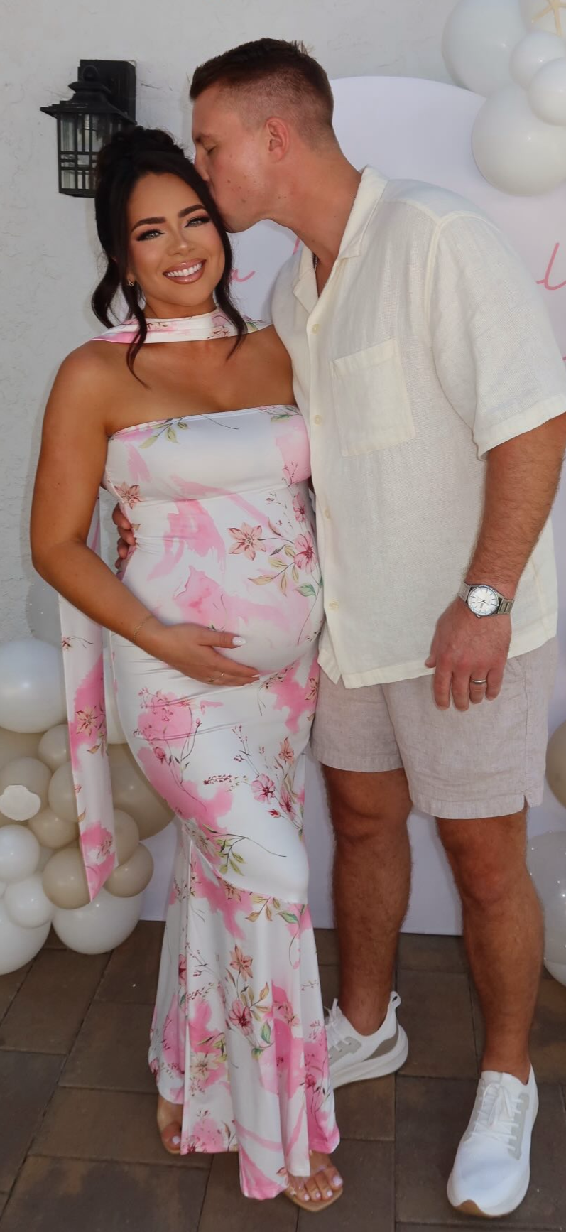 A pregnant woman smiling and a man with his eyes closed, kissing her forehead at a celebration with balloons.