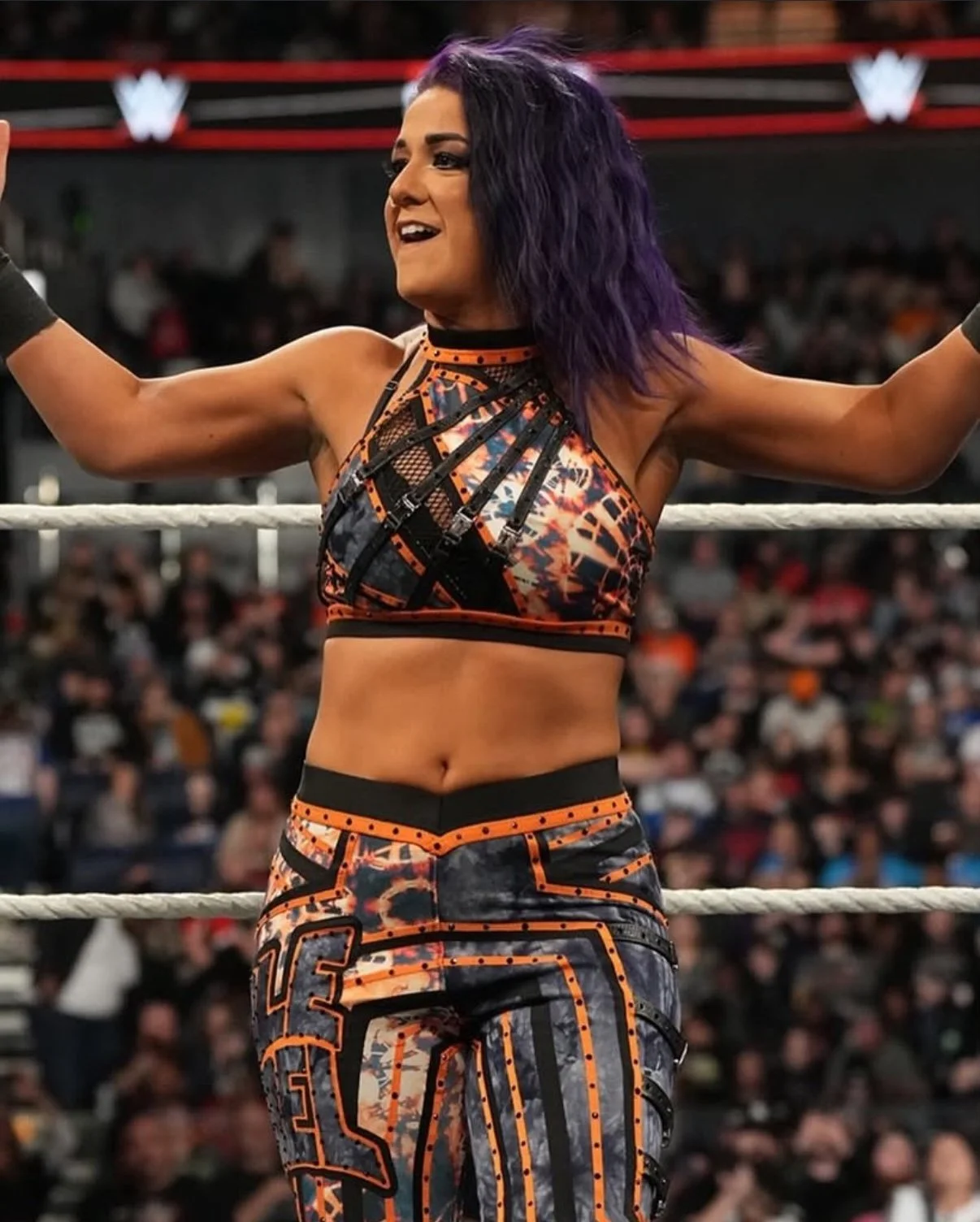 A professional wrestler standing in the wrestling ring with her arms raised, wearing a colorful, patterned sports bra and matching pants, with purple hair and a confident expression. WWE