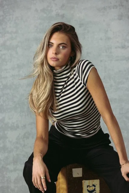 A young woman with long, wavy blonde hair and blue eyes wearing a black and white striped t-shirt, sitting on a wooden box against a gray textured background.