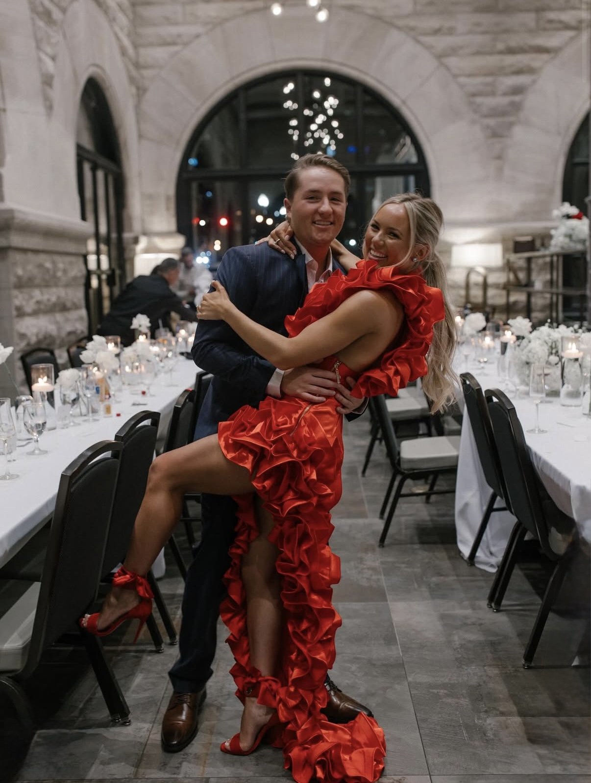 A man and woman dancing and smiling at an indoor event, with decorated tables and dim lighting.