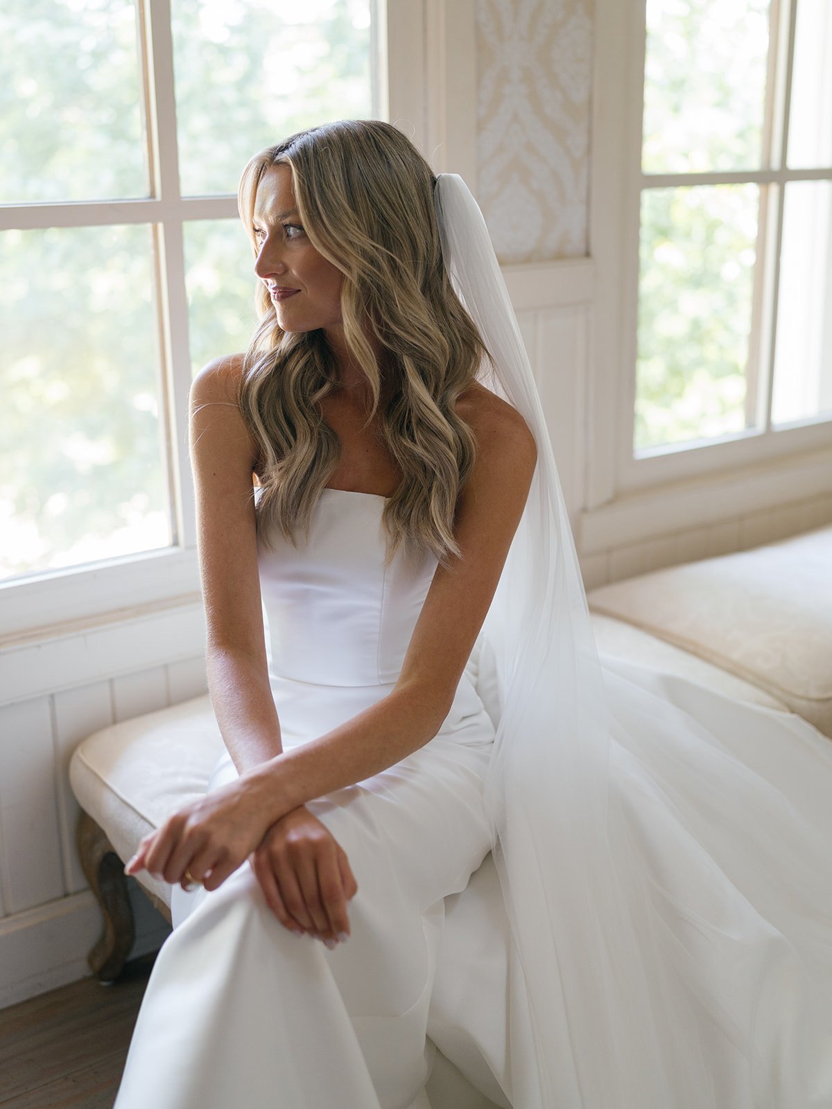 Bride sitting on a window seat, wearing a strapless white wedding gown with a veil, looking out the window with a serene expression.